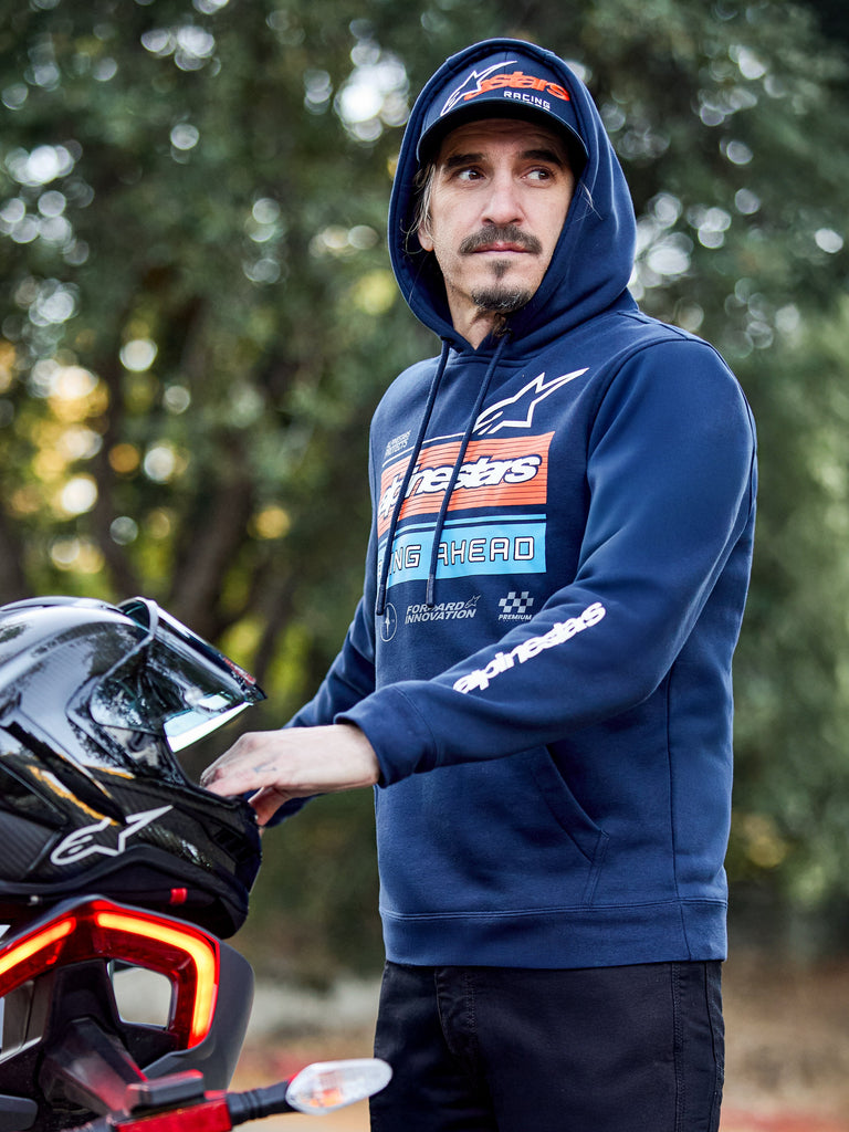 Alpinestars Harken Hoodie, Navy Blue, pullover fleece with gradation fade chest graphic and white Alpinestars wordmark on sleeve, worn by a man holding a motorcycle helmet, casual motorsport-inspired streetwear