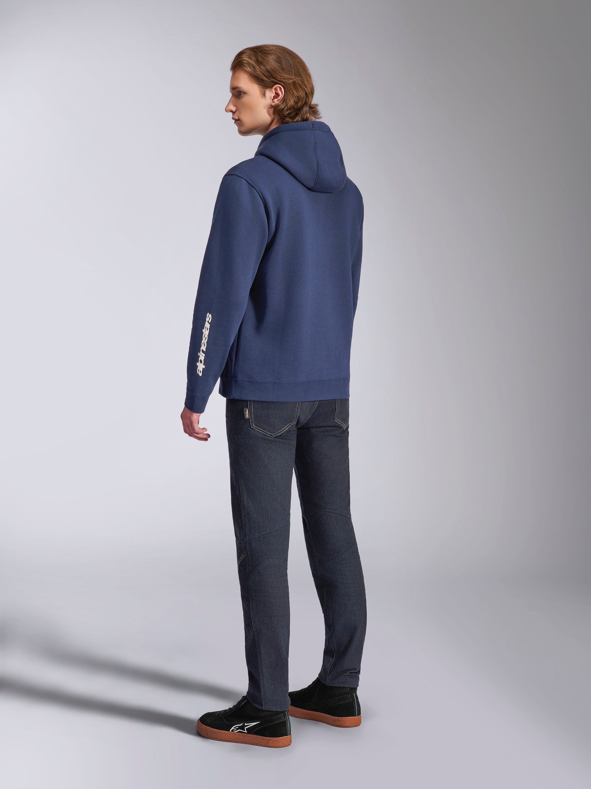Alpinestars Harken Hoodie in Navy, back view of male model, casual pullover fleece sweatshirt with white wordmark logo on the sleeve, paired with dark denim jeans and black sneakers, modern motorsport-inspired streetwear