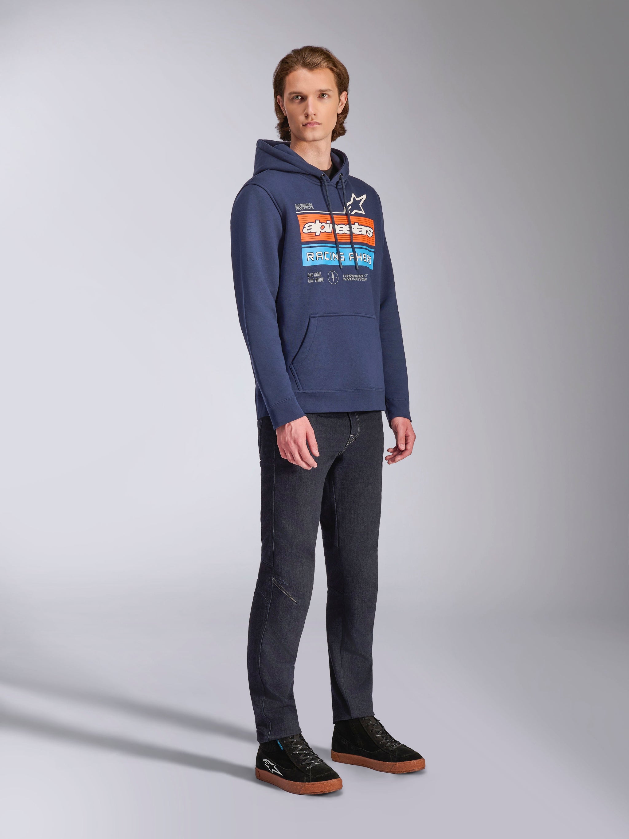 Alpinestars Harken Hoodie in Navy Blue, worn by a male model with dark denim pants and black sneakers, featuring a large chest graphic with orange and light blue accents, Alpinestars wordmark, and kangaroo pocket.