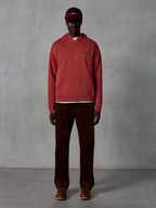 Alpinestars Essentials Ageless Hoodie, casual sportswear hoodie in Brick red with a garment stone wash finish, featuring a small black Alpinestars chest logo and front pouch pocket, worn by a model with a maroon cap and brown corduroy pants for a clean streetwear look.