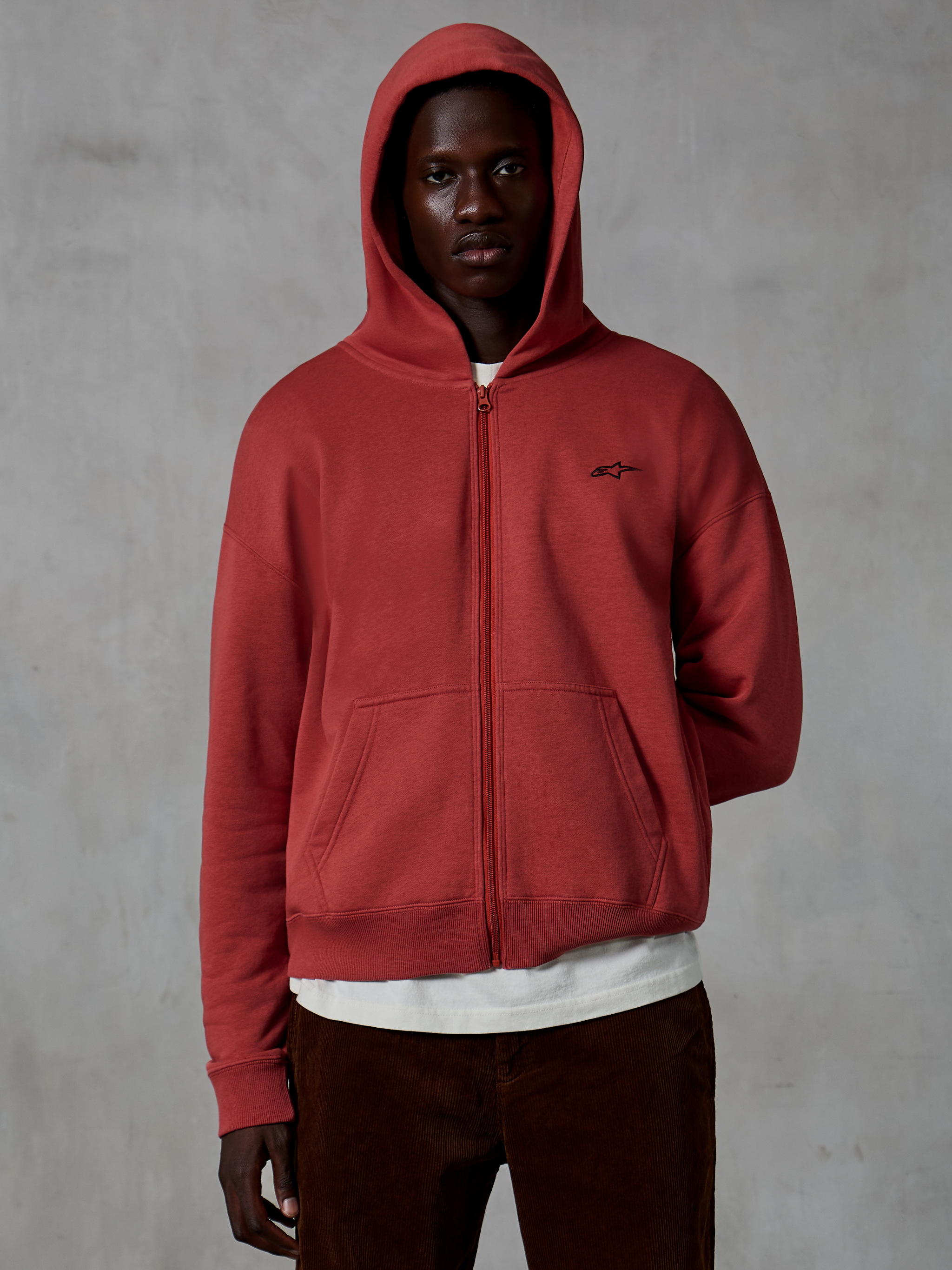 Alpinestars Essentials Ageless Zip Hoodie, casual sportswear in Brick red, full-zip design with hood up, featuring a small black logo on the chest, worn by a model with a white t-shirt and brown corduroy pants against a gray background.