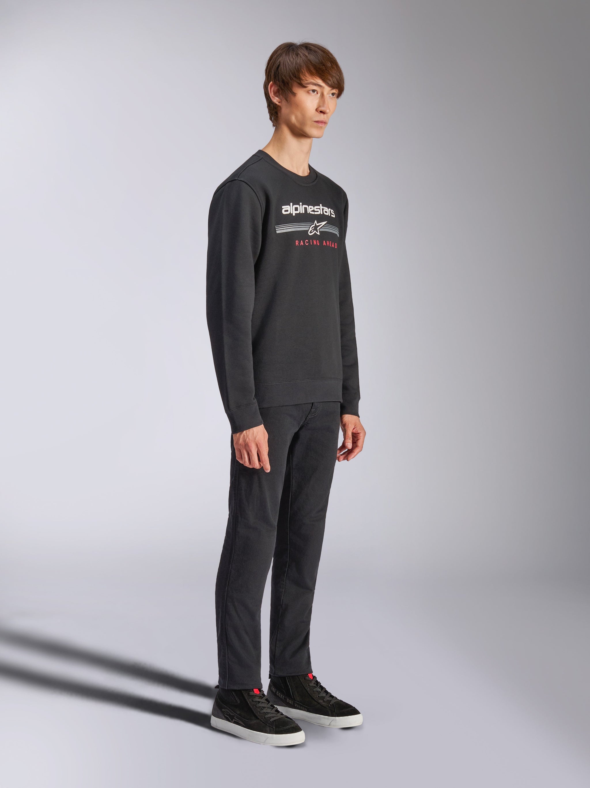 Alpinestars Bettern Crew Sweatshirt, black crewneck pullover, white alpinestars logo with 'RACING AHEAD' in red on the chest, paired with black pants and sneakers, casual motorsport-inspired streetwear