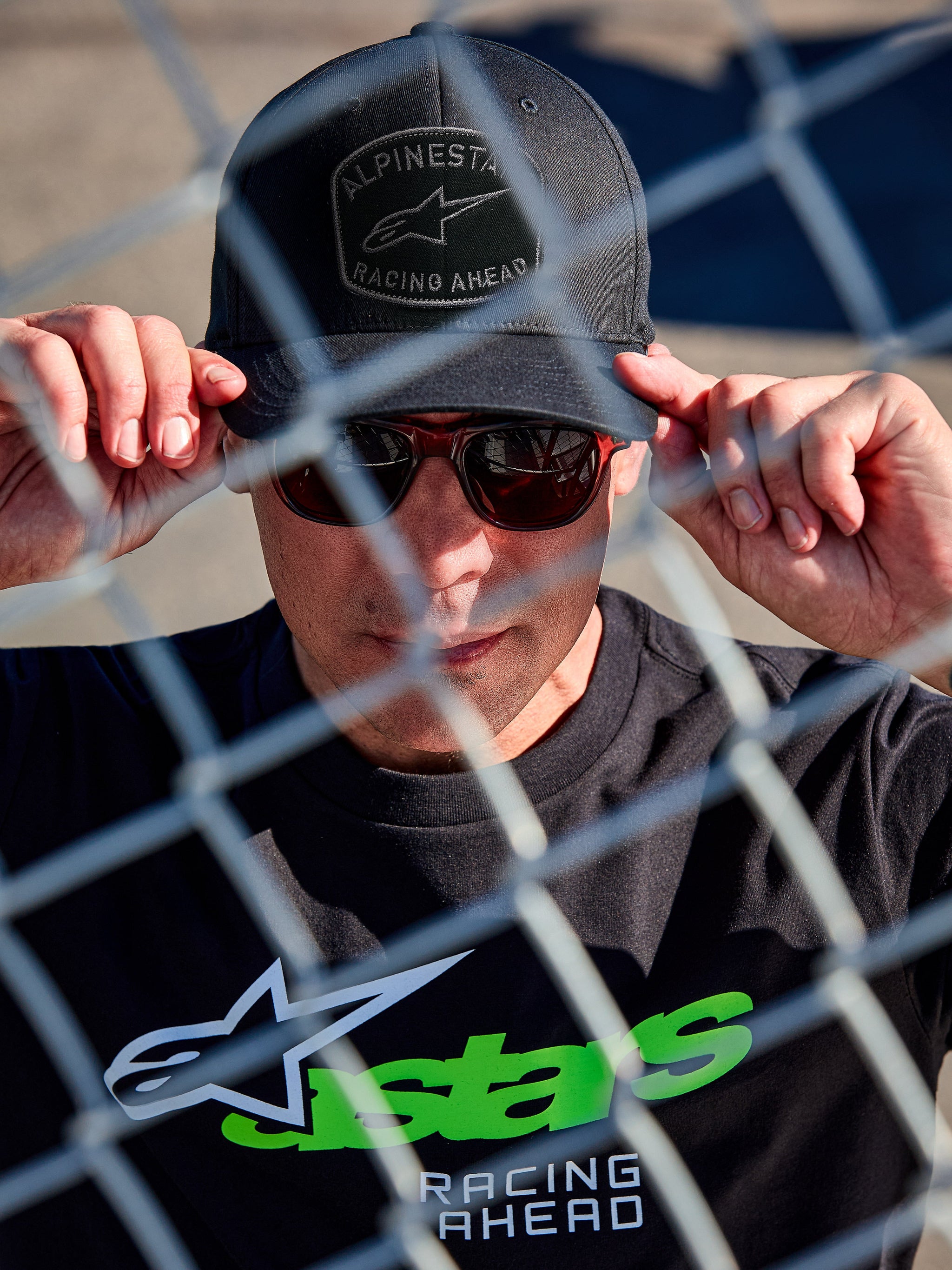 Alpinestars Promptus Snapback Hat, black lifestyle cap, flat bill with structured crown, front curved embroidery patch with braided rope, worn by a man with sunglasses and black t-shirt with green Astars logo, shot through a chain-link fence