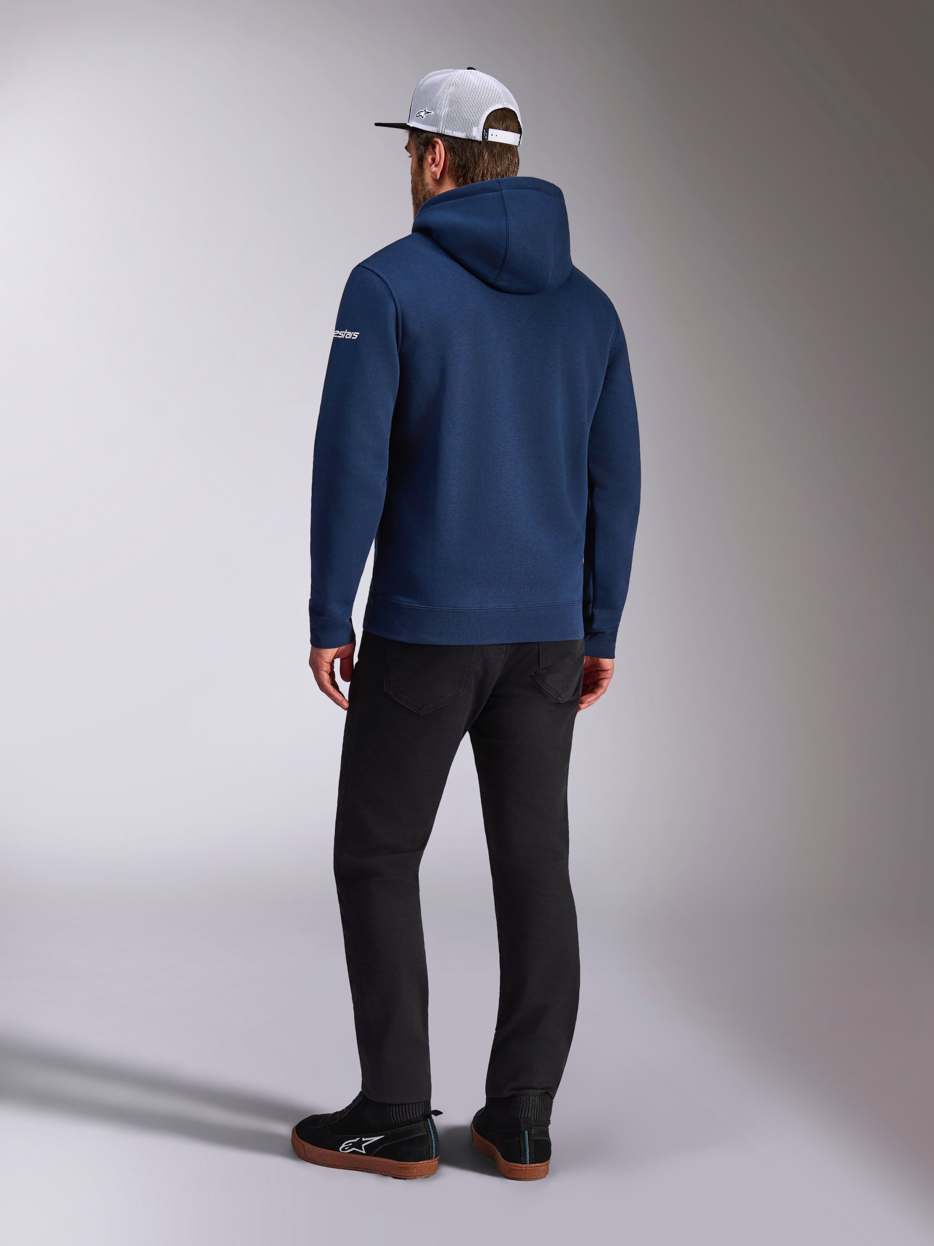 Alpinestars Frontal Hoodie, Rear view of a navy blue pullover fleece hoodie, white word logo on the sleeve, paired with black pants and a white mesh trucker hat, casual motorsport-inspired streetwear