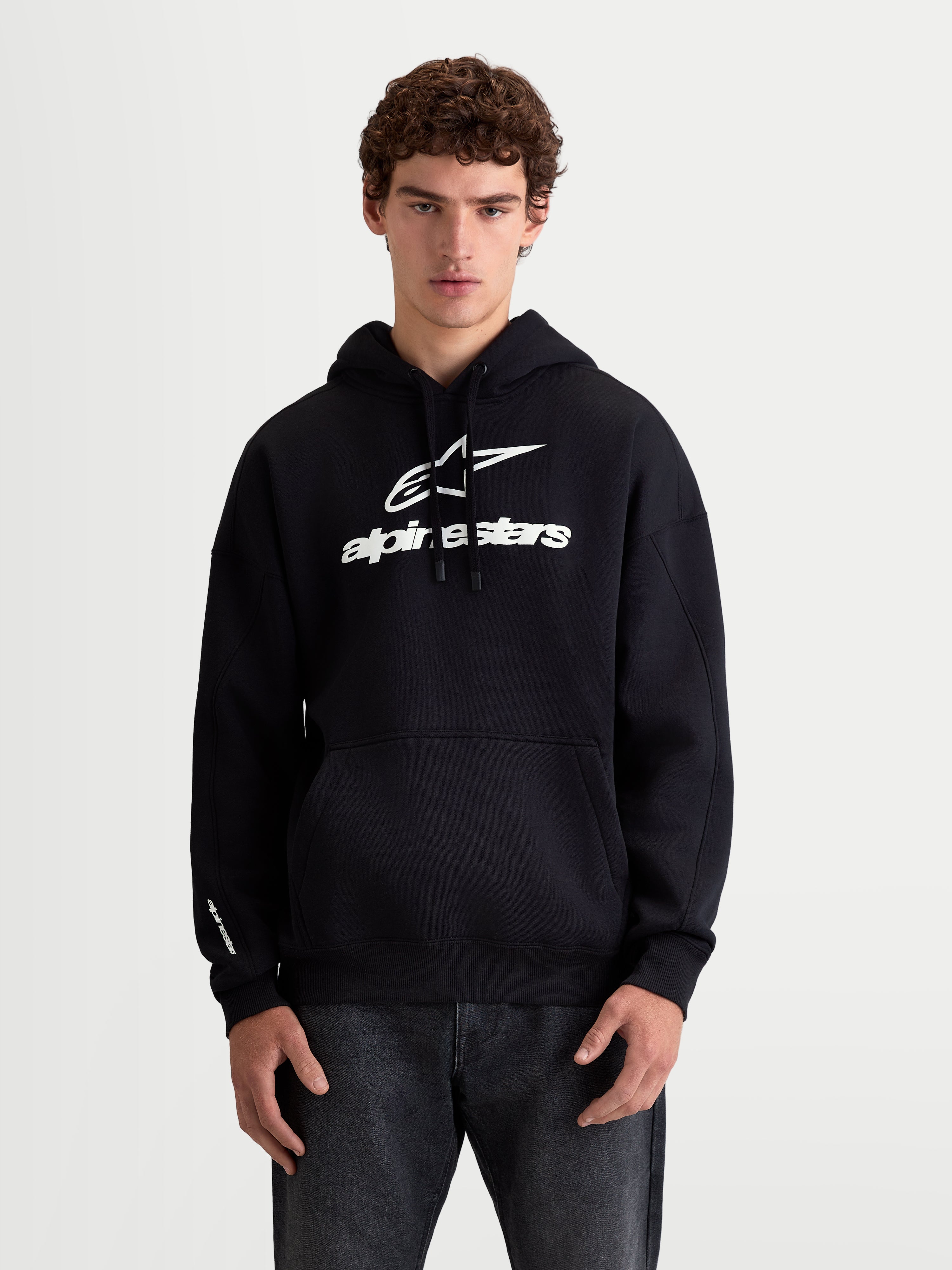 Alpinestars Prelude Oversized Hoodie, Men's and Women's Sportswear Fleece Pullover on model, Black and White, Cotton and Recycled Polyester. Features a relaxed oversized fit, bold white puff-print logos on the chest and sleeve, kangaroo pocket, and adjustable hood for daily casual wear.
