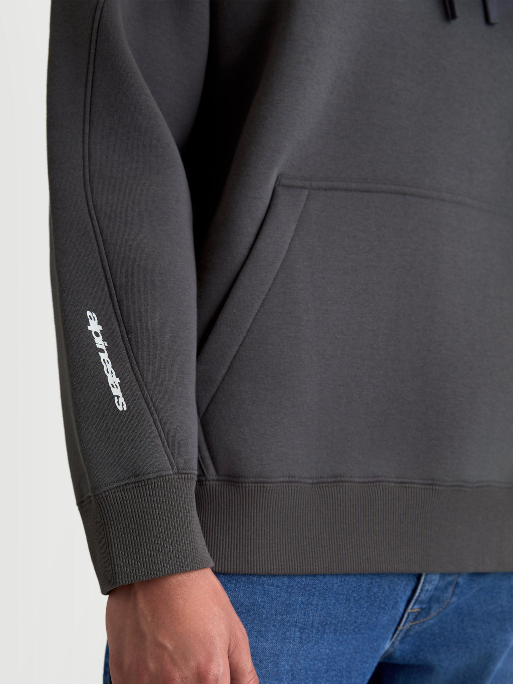 Alpinestars Prelude Oversized Hoodie, Sportswear Fleece Pullover in Charcoal Gray, detail shot showing the white puff-print logo on the sleeve, decorative seaming, and ribbed cuff and hem.
