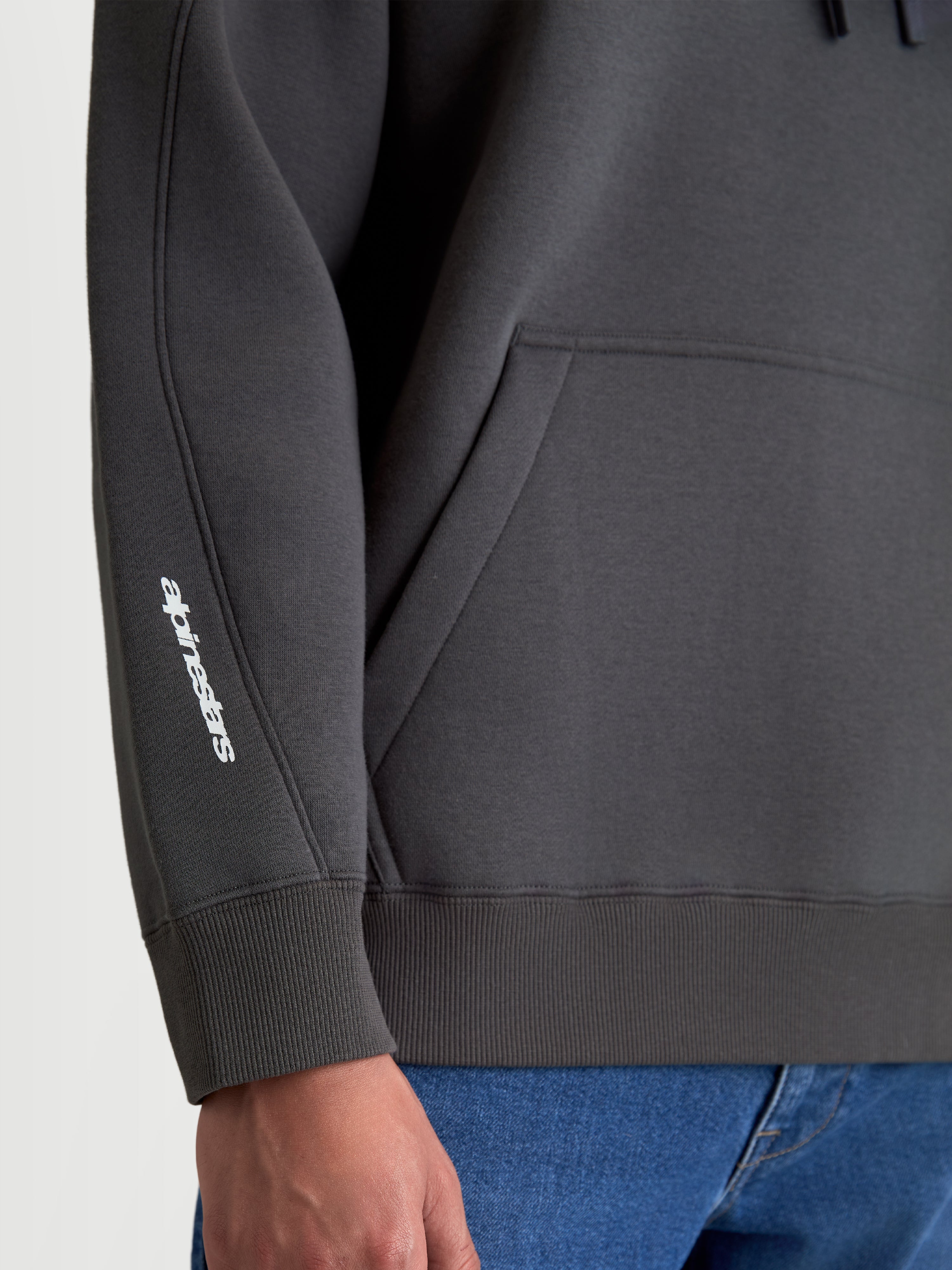 Alpinestars Prelude Oversized Hoodie, Sportswear Fleece Pullover in Charcoal Gray, detail shot showing the white puff-print logo on the sleeve, decorative seaming, and ribbed cuff and hem.