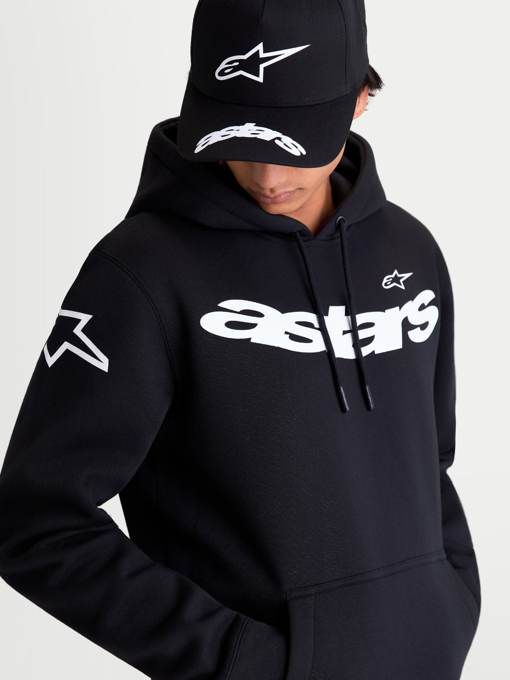 Alpinestars Velocifier Hoodie, Black Sportswear Pullover Fleece, on-model view showcasing the white curved Astars wordmark on the chest and star logo on the shoulder. This premium casual hoodie features a drawstring hood and kangaroo pocket, ideal for off-bike motorsports style.