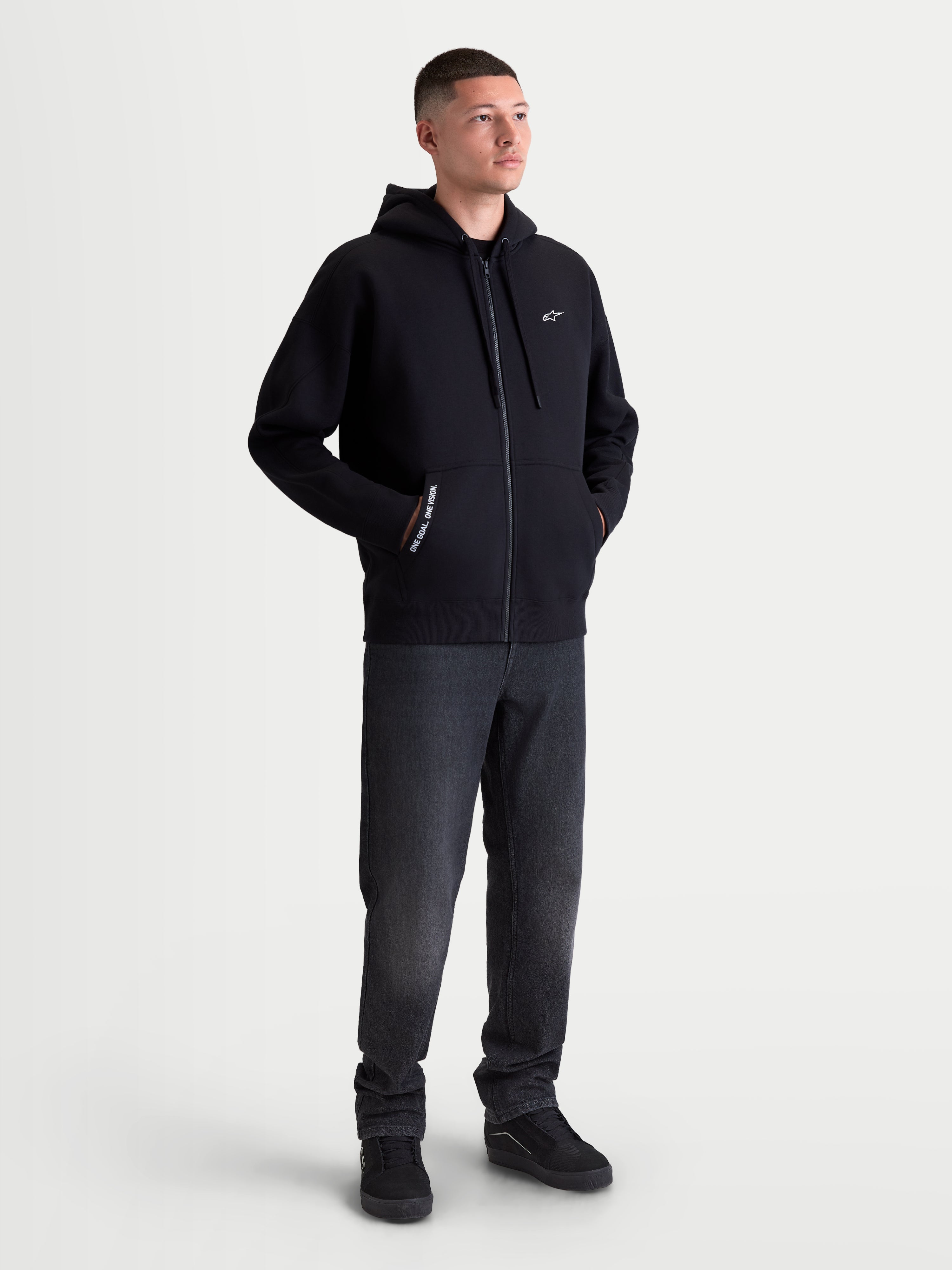 Alpinestars Pioneer Oversized Hoodie, Sportswear Full-Zip Fleece for Men and Women, Black, Cotton-Rich Blend, featuring a Vislon zipper, kangaroo pocket with 'ONE GOAL. ONE VISION.' branding, and white Astars chest logo. On-model view showing the relaxed oversized fit designed for casual everyday wear.