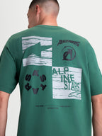 Alpinestars Checkback Oversized CSF Tee, Green Sportswear Short Sleeve T-Shirt, Men's and Women's Casual Wear, Back View on Model. Features a prominent black and white back checkerboard graphic with Astars logos and 'One Goal One Vision' text. Constructed from a soft cotton-poly blend with an oversized fit for comfort and motorsport-inspired style.