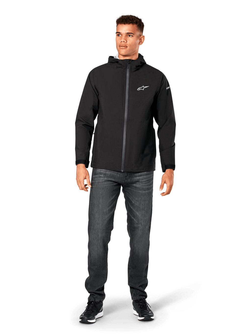 Alpinestars Kascade Rain Jacket, Men's Rain Shell, Black, worn with dark grey jeans and black sneakers, featuring a hooded design, coated front zipper, and white Alpinestars logo on the chest, casual motorsport-inspired streetwear