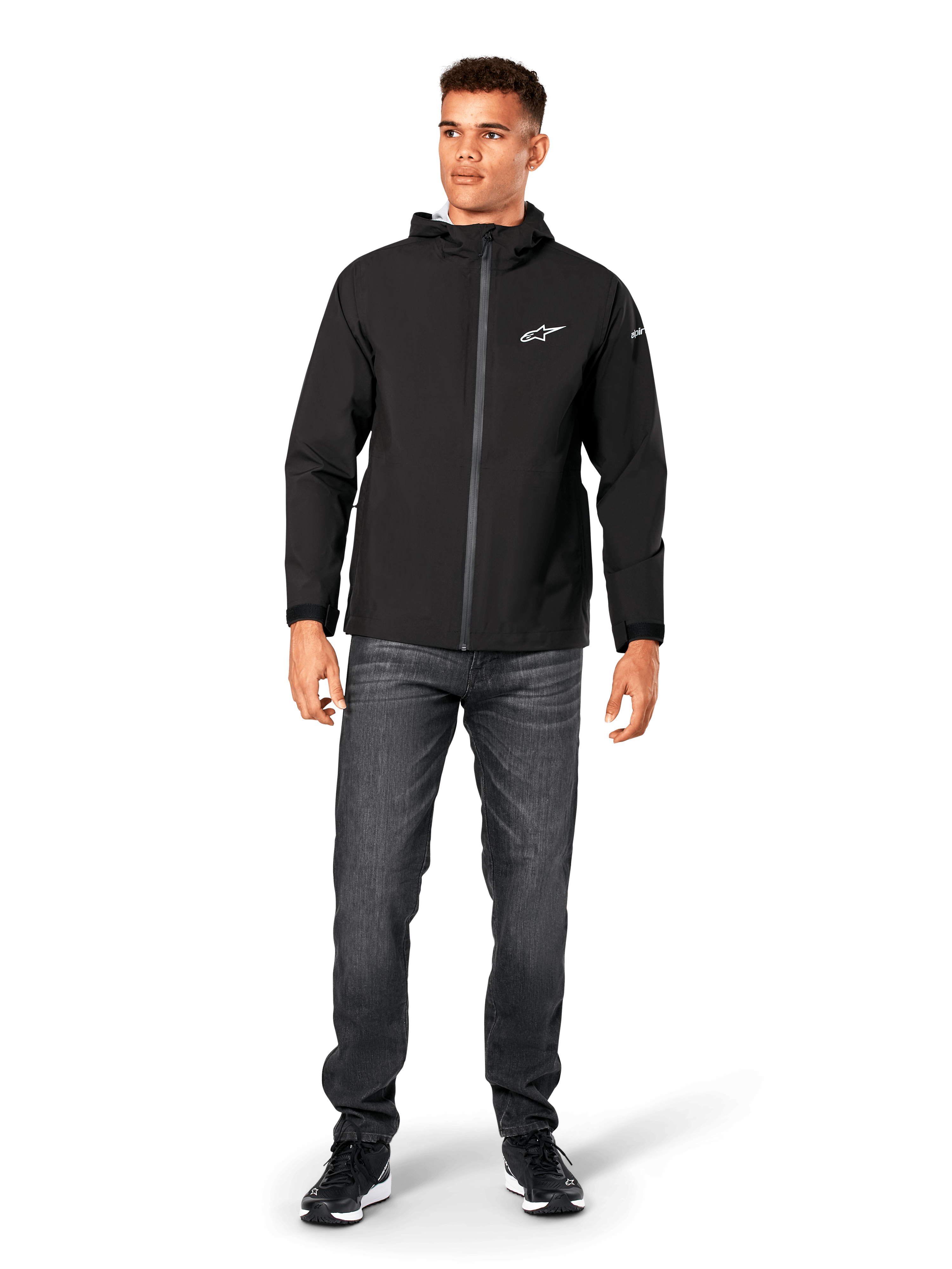 Alpinestars Kascade Rain Jacket, Men's Rain Shell, Black, worn with dark grey jeans and black sneakers, featuring a hooded design, coated front zipper, and white Alpinestars logo on the chest, casual motorsport-inspired streetwear