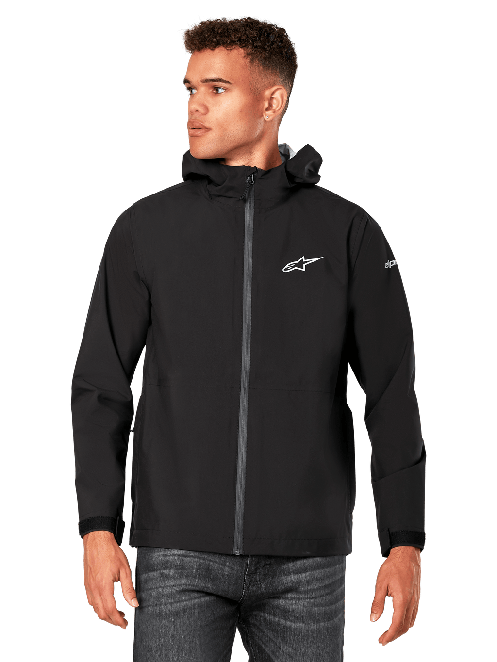 Male model wearing Alpinestars Kascade Rain Jacket, Black, white chest logo and sleeve branding, hooded rain shell with coated front zipper and velcro cuffs, paired with dark jeans, casual motorsport-inspired streetwear