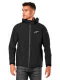 Male model wearing Alpinestars Kascade Rain Jacket, Black, white chest logo and sleeve branding, hooded rain shell with coated front zipper and velcro cuffs, paired with dark jeans, casual motorsport-inspired streetwear