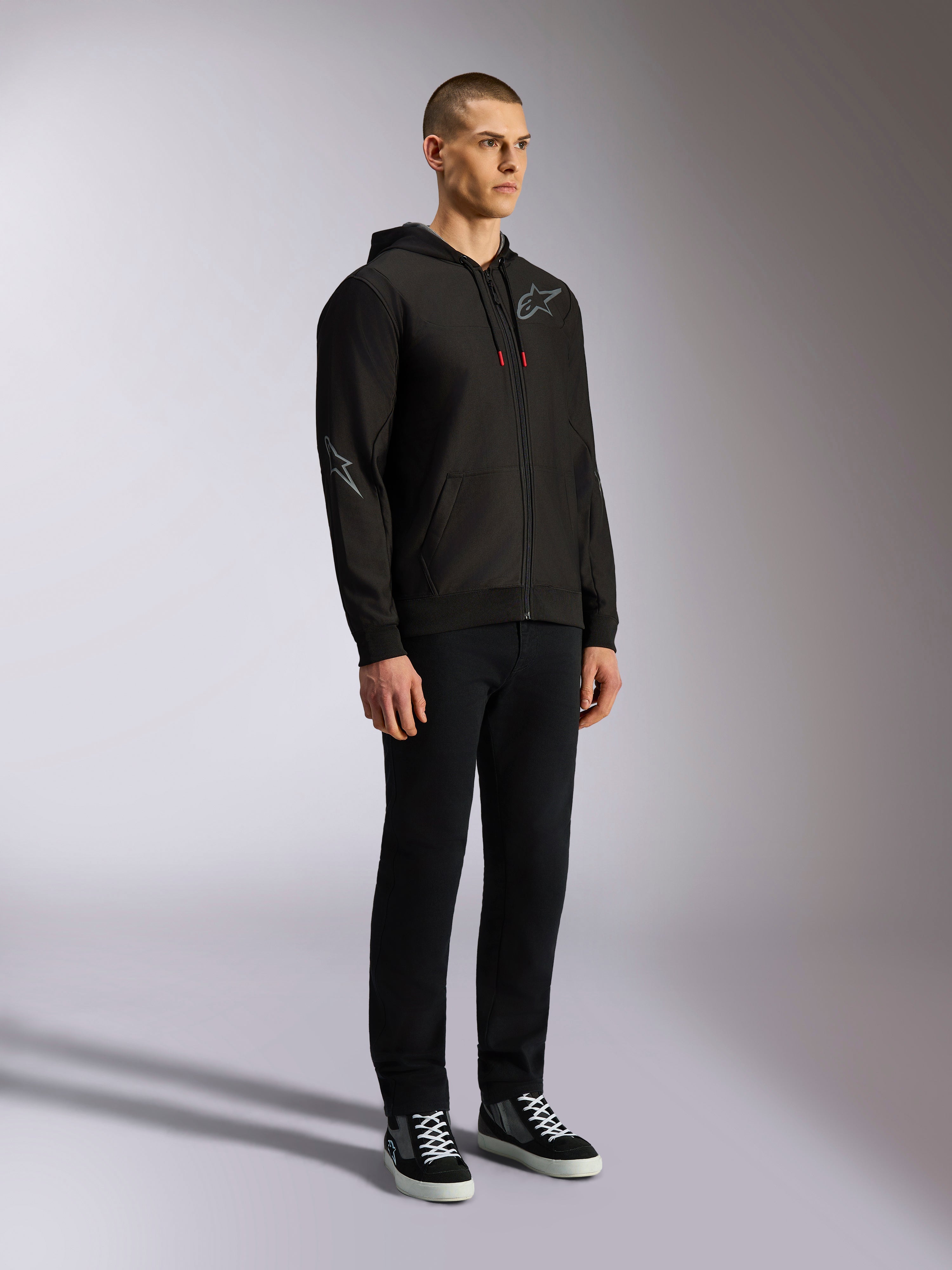 Alpinestars Sessions V3 Hoodie, casual motorsport streetwear, black zip-up hoodie with gray star logo on chest and sleeve, red-tipped drawcords, rib cuffs and hem, worn by model with black pants and sneakers.