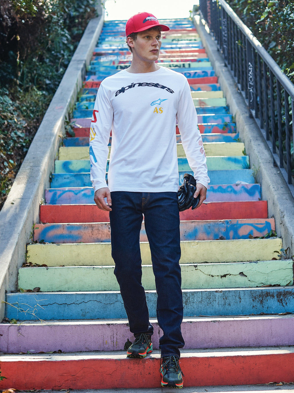 Alpinestars Brundo CSF Long Sleeve Tee, white t-shirt with black Alpinestars logo and colorful graphic prints, worn by a model on colorful stairs, paired with dark blue jeans and a red cap, casual motorsport streetwear.