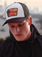 Alpinestars Hardware Trucker Hat in Military White, worn by a man in a black hoodie, featuring a white front panel with a black curved bill and black mesh back, tan embroidered patch with red Alpinestars Motocross branding, casual lifestyle motorsport streetwear