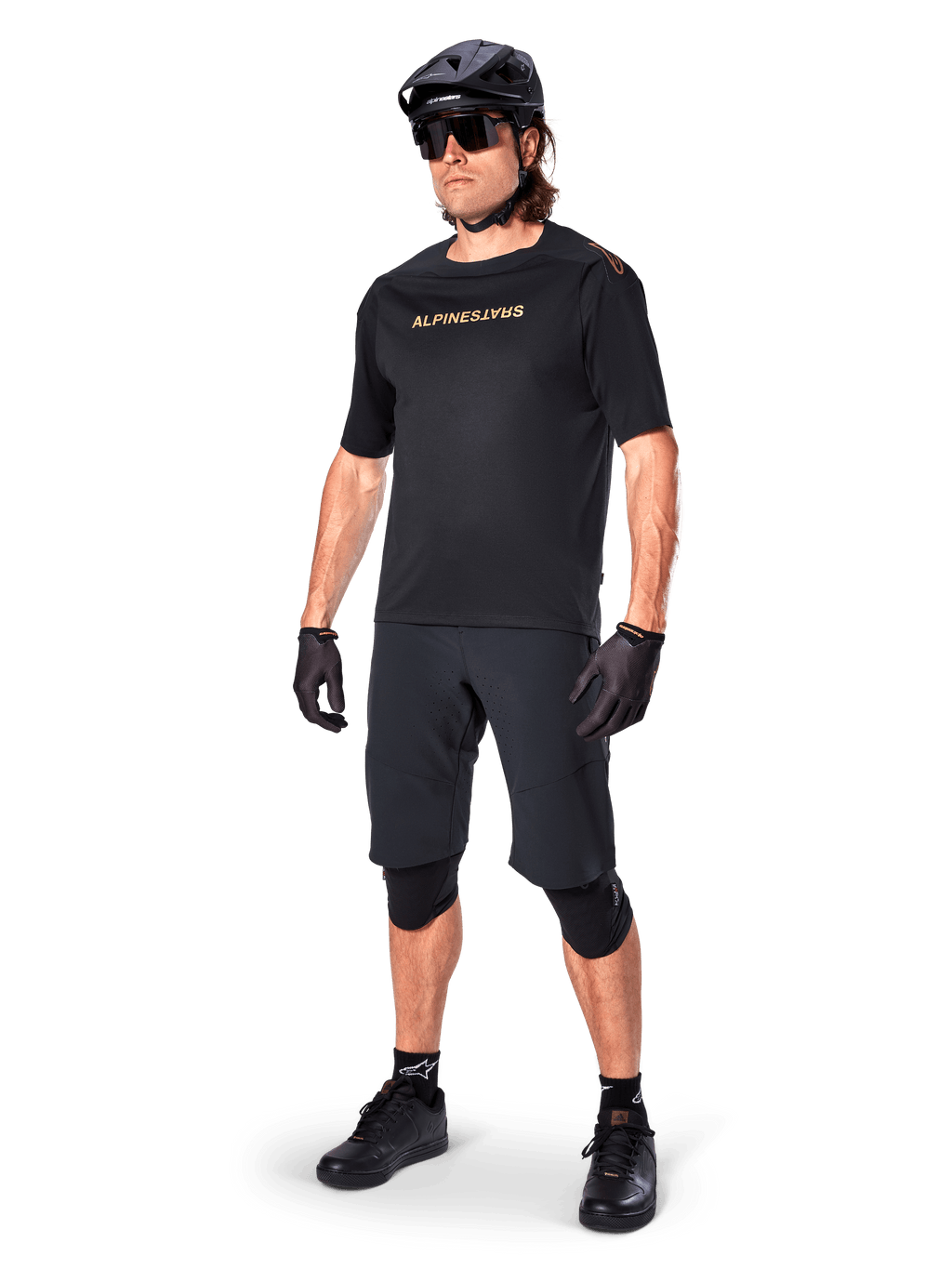 Cyclist wearing Alpinestars A-Aria Elite Shorts, Black Mountain Bike Shorts, paired with a matching black jersey with gold branding, black helmet, sunglasses, and knee pads, complete performance outfit for off-road cycling