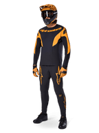 Mountain bike rider wearing complete racing gear, Alpinestars A-Supra Race Pants in black with orange accents, matching black and orange long-sleeve jersey, orange helmet with black goggles, orange and black gloves, and black shoes, professional MTB racing kit design