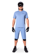 Model wearing Alpinestars A-Dura Shorts in Infinity Blue, matching light blue jersey, black mountain bike helmet, black sunglasses, black gloves and knee pads, complete MTB outfit for trail riding