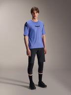 Male model wearing Alpinestars A-Aria Lite Shorts in black, paired with a blue A-Aria Polartec Elite Short Sleeve Jersey, black knee guards, and black cycling shoes, performance mountain bike apparel for trail riding.
