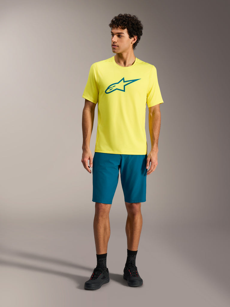 Yellow Alpinestars A-Dura Ride SS Jersey and teal-green shorts, mountain bike performance apparel in a studio setting.