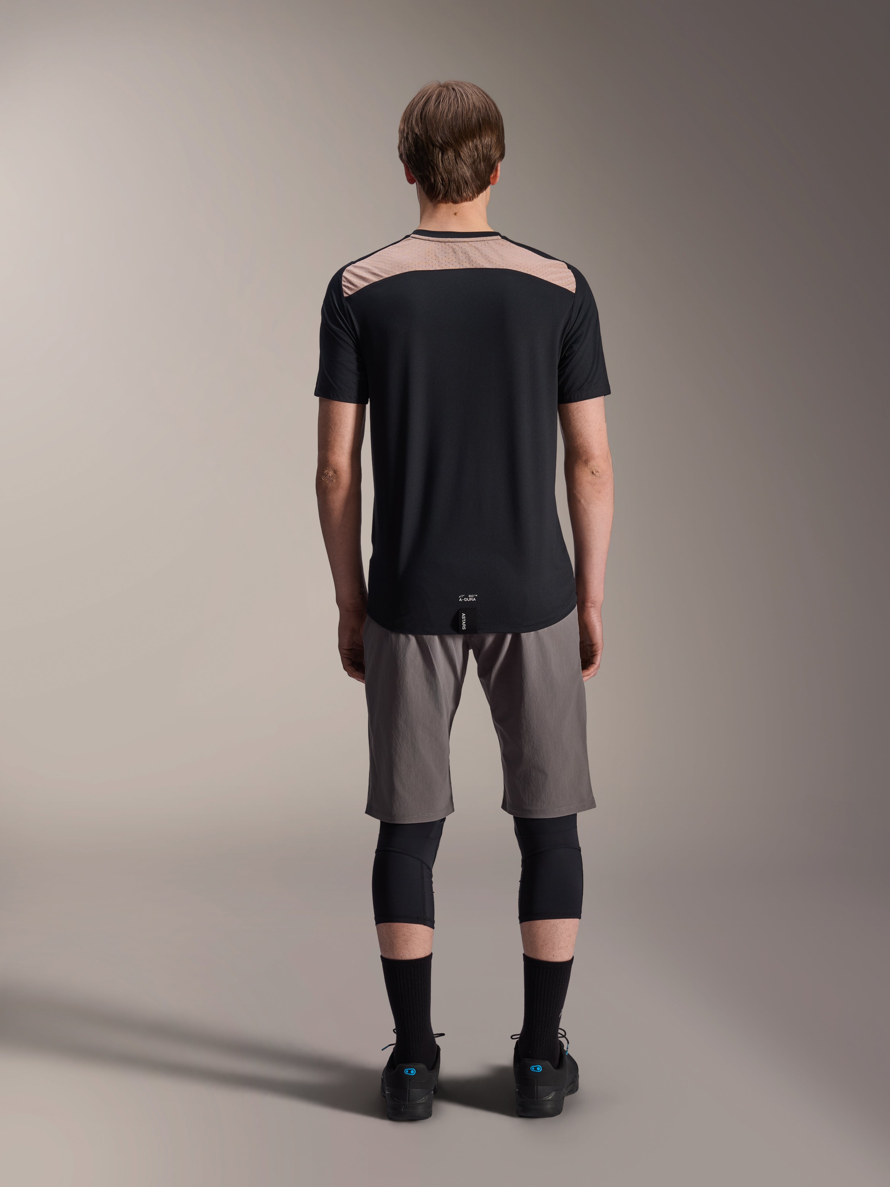 Alpinestars A-Dura Lipan Jersey - Short Sleeve, Black with tan upper back panel, rear view showing dropped tail, worn with grey MTB shorts and black knee guards, performance mountain bike apparel