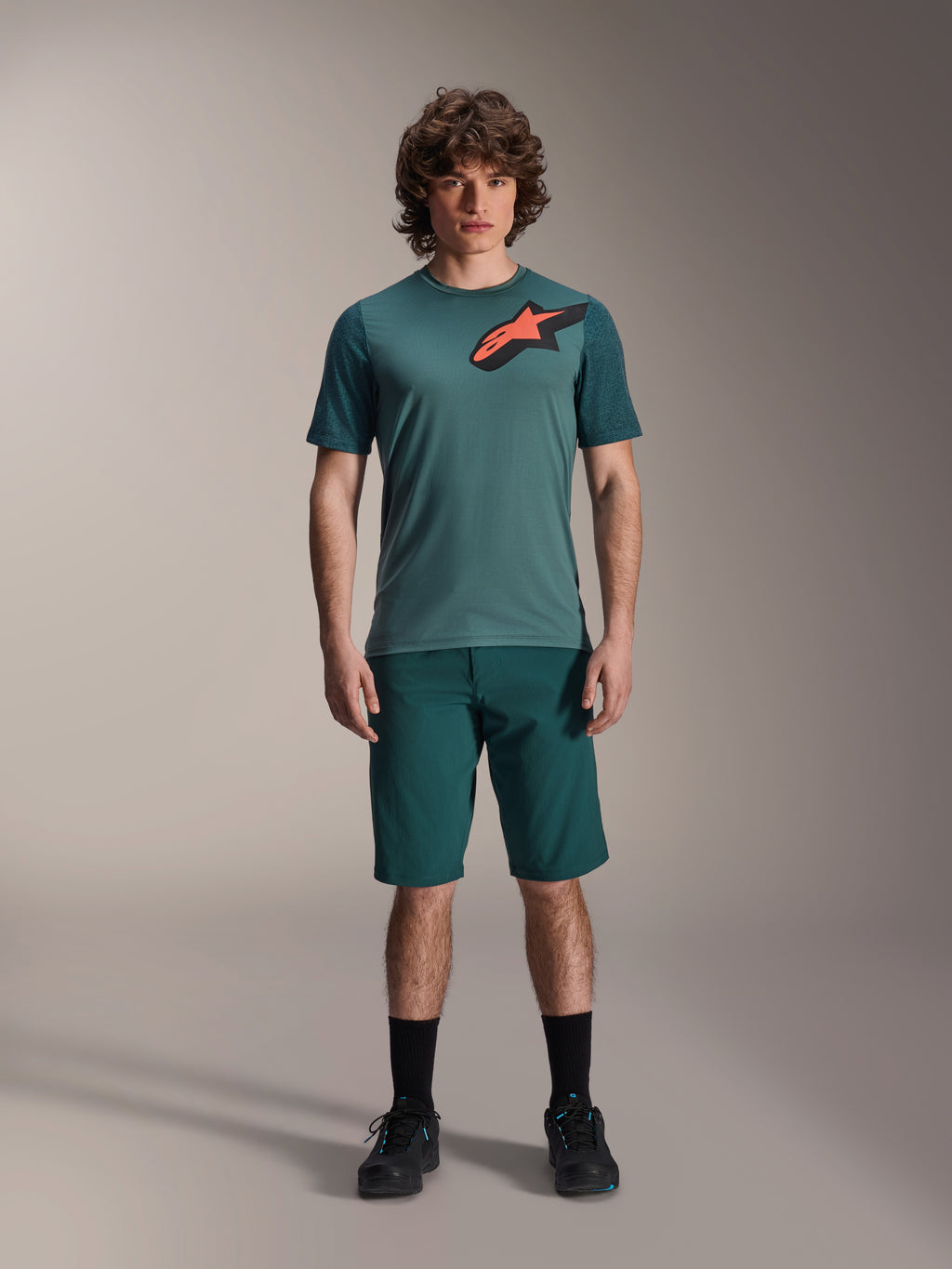 Alpinestars A-Dura Shorts in Dark Pine, men's mountain bike shorts, shown on a model with a matching teal short-sleeve jersey featuring a red and black star logo, black socks and cycling shoes, casual sporty mountain biking apparel