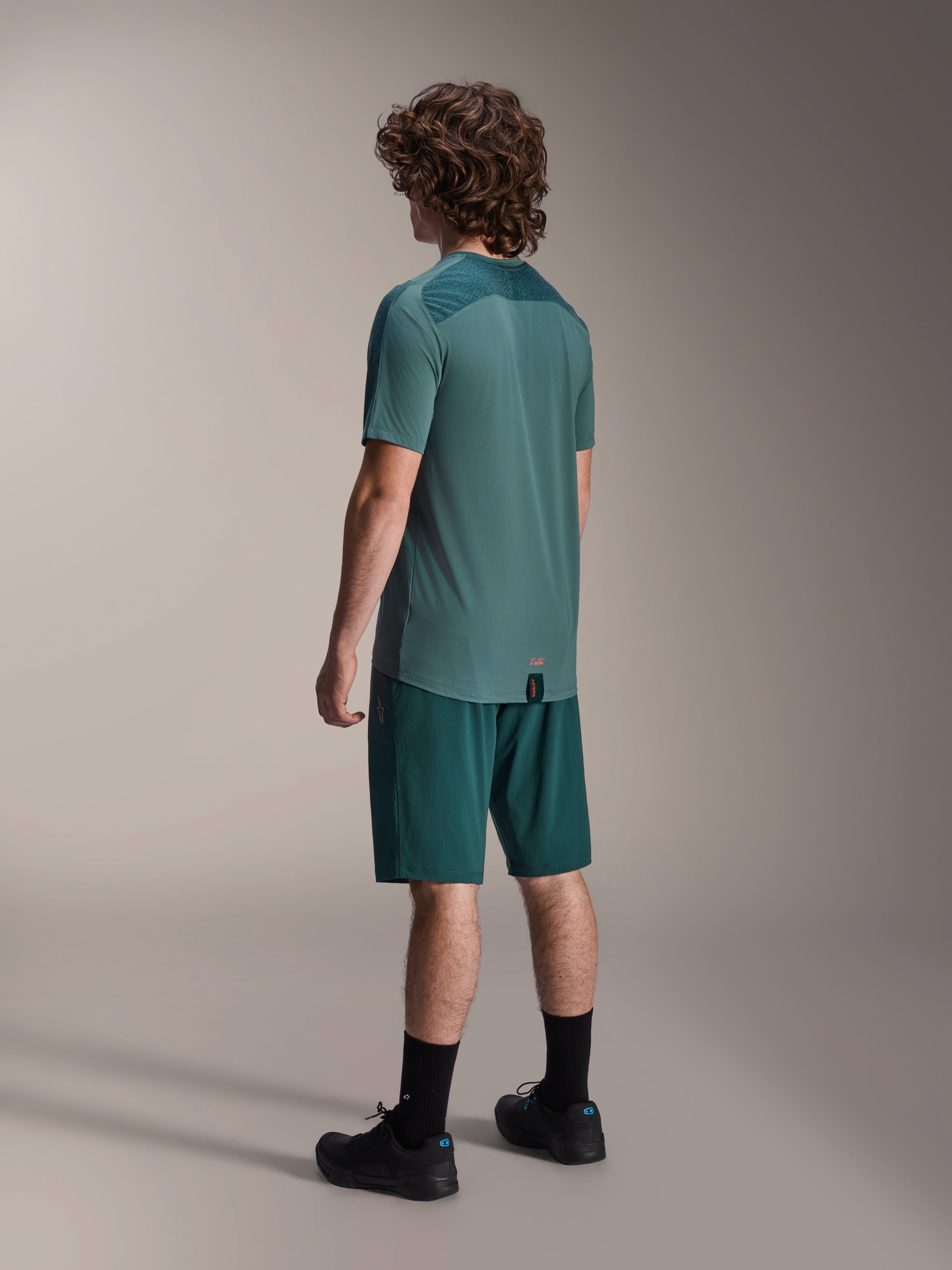 Model wearing Alpinestars A-Dura Shorts in Dark Pine and matching green Lipan short-sleeve jersey, back view, paired with black socks and black mountain bike shoes, professional MTB riding gear.