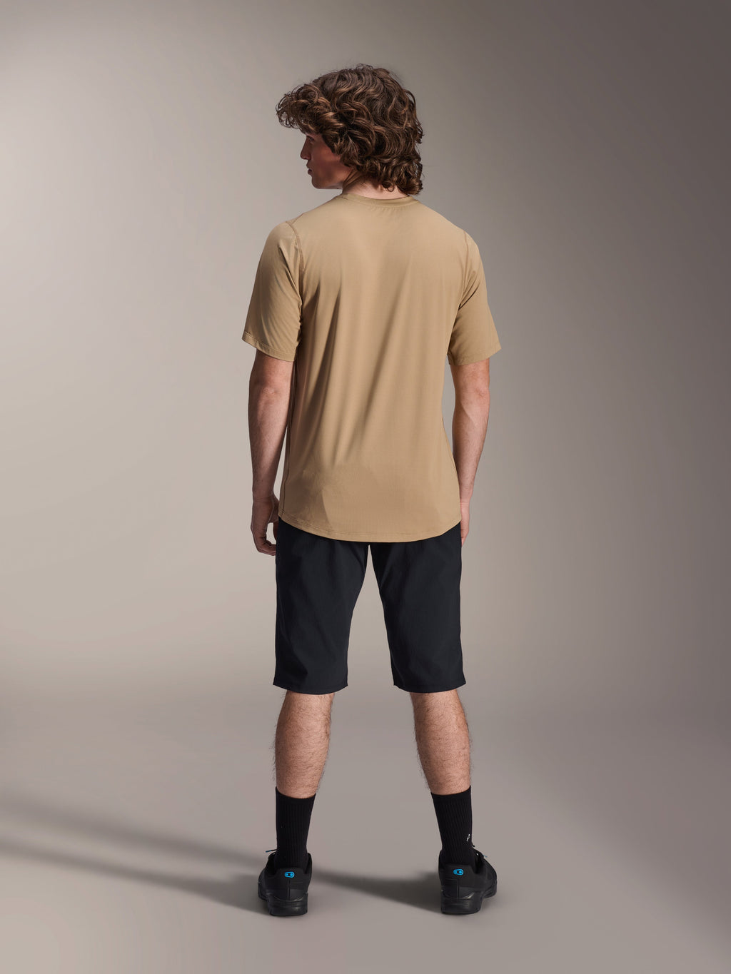 Male rider seen from the back wearing the Alpinestars A-Dura Astar Short Sleeve Jersey in Light Brown, paired with black mountain bike shorts and black socks, casual MTB trail riding style.
