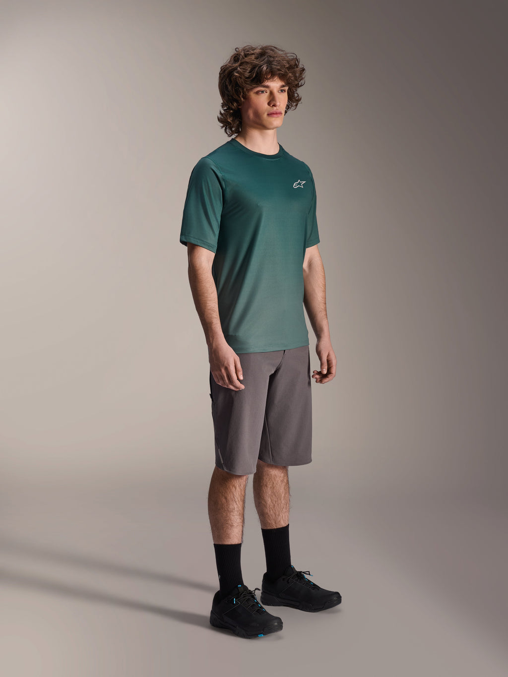 Alpinestars A-Dura Rise Jersey - Short Sleeve in Silver Pine, green mountain bike jersey worn by a model with grey shorts and black sneakers, casual MTB trail riding gear featuring a small white chest logo
