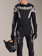 Alpinestars A-Supra Melt MTB Racing Kit, Long Sleeve Jersey and Pants, Black and Grey with white Astars logos, model holding a black full-face helmet and wearing matching gloves, high-performance gear for downhill and enduro mountain biking