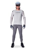 Alpinestars A-Aria Polartec Switch Jersey, Long Sleeve, Light Gray, worn by a male mountain bike rider with dark gray MTB pants, light blue helmet, and sunglasses, featuring chest branding and laser-perforated ventilation on the sleeves, complete off-road riding outfit.