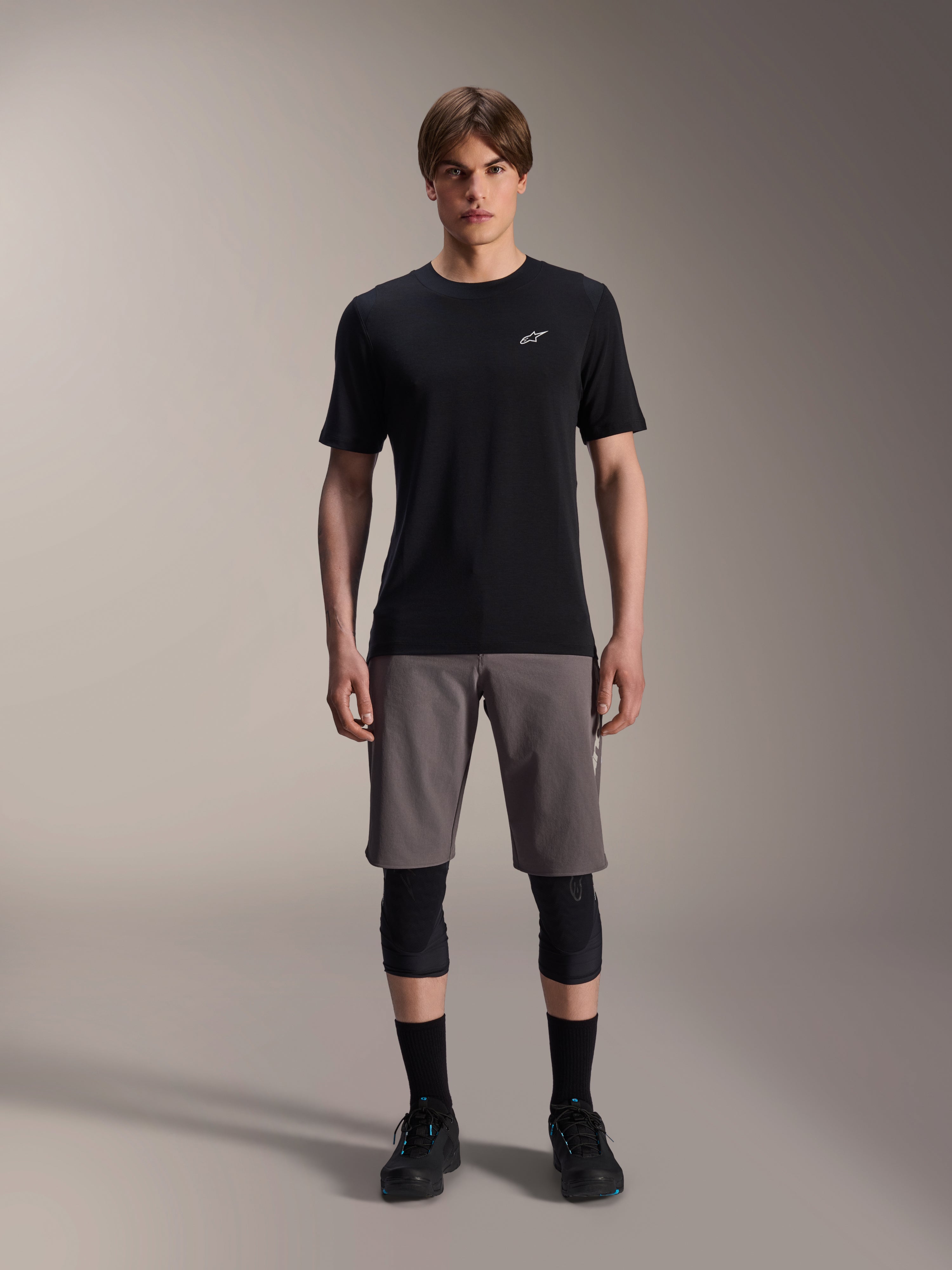 Male model wearing Alpinestars A-Dura Elite Shorts in Dark Gray and A-Dura Dri-Wool Short Sleeve Jersey in Black, paired with black knee guards and cycling shoes, technical mountain bike apparel for off-road performance.