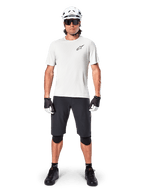 Male cyclist wearing Alpinestars A-Aria Astar Short Sleeve Jersey in White Dove, featuring a black Astars logo on the chest, paired with black mountain bike shorts, white helmet, sunglasses, black knee guards, and gloves, complete off-road cycling outfit.