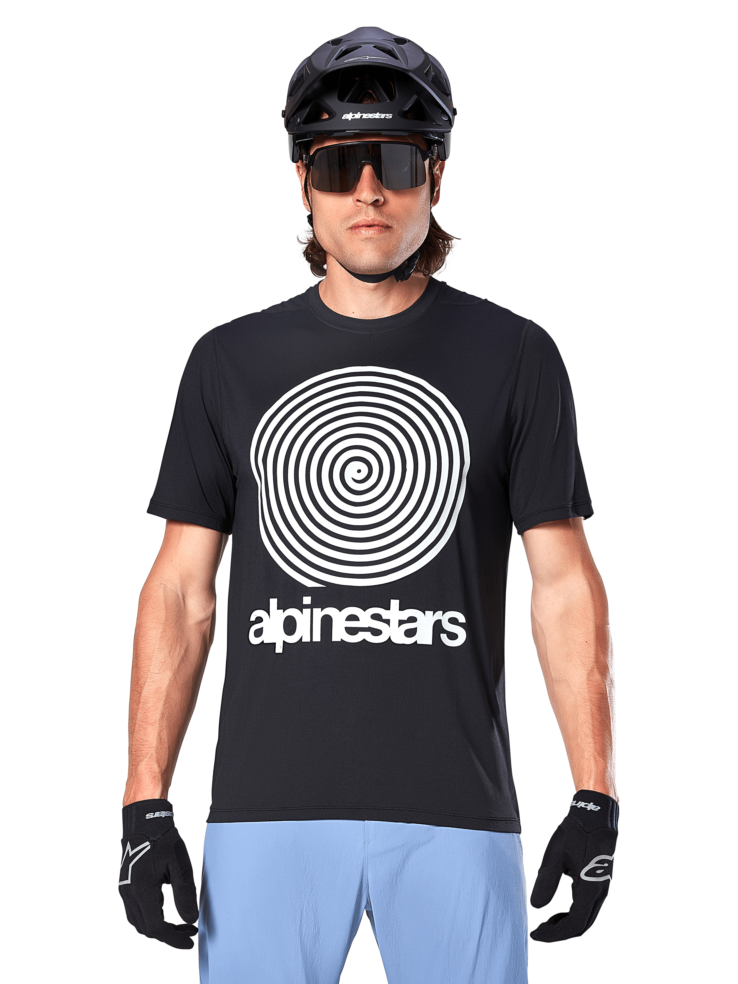 Alpinestars A-Dura Oscar Jersey, Short Sleeve MTB Jersey, Black, featuring a large white vintage Oscar spiral graphic on the chest, worn with a black cycling helmet, sunglasses, black gloves, and light blue shorts, heritage-inspired off-road cycling gear