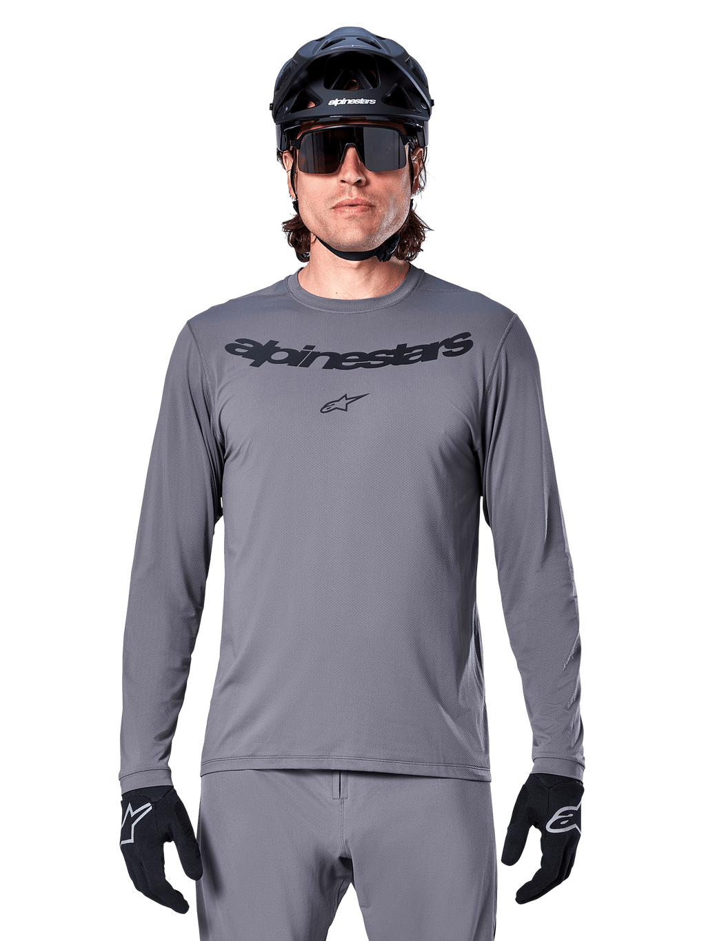 Male rider wearing the Alpinestars A-Dura Rocker Long Sleeve Jersey, Dark Gray MTB jersey with black wordmark on chest, paired with gray pants, black helmet, and gloves, designed for off-road cycling