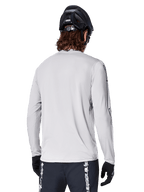Alpinestars A-Dura Switch Long Sleeve Jersey, Light Gray, back view of a mountain biker wearing a black MTB helmet and gloves, featuring a drop tail for extra coverage and textured jacquard fabric, performance cycling gear.