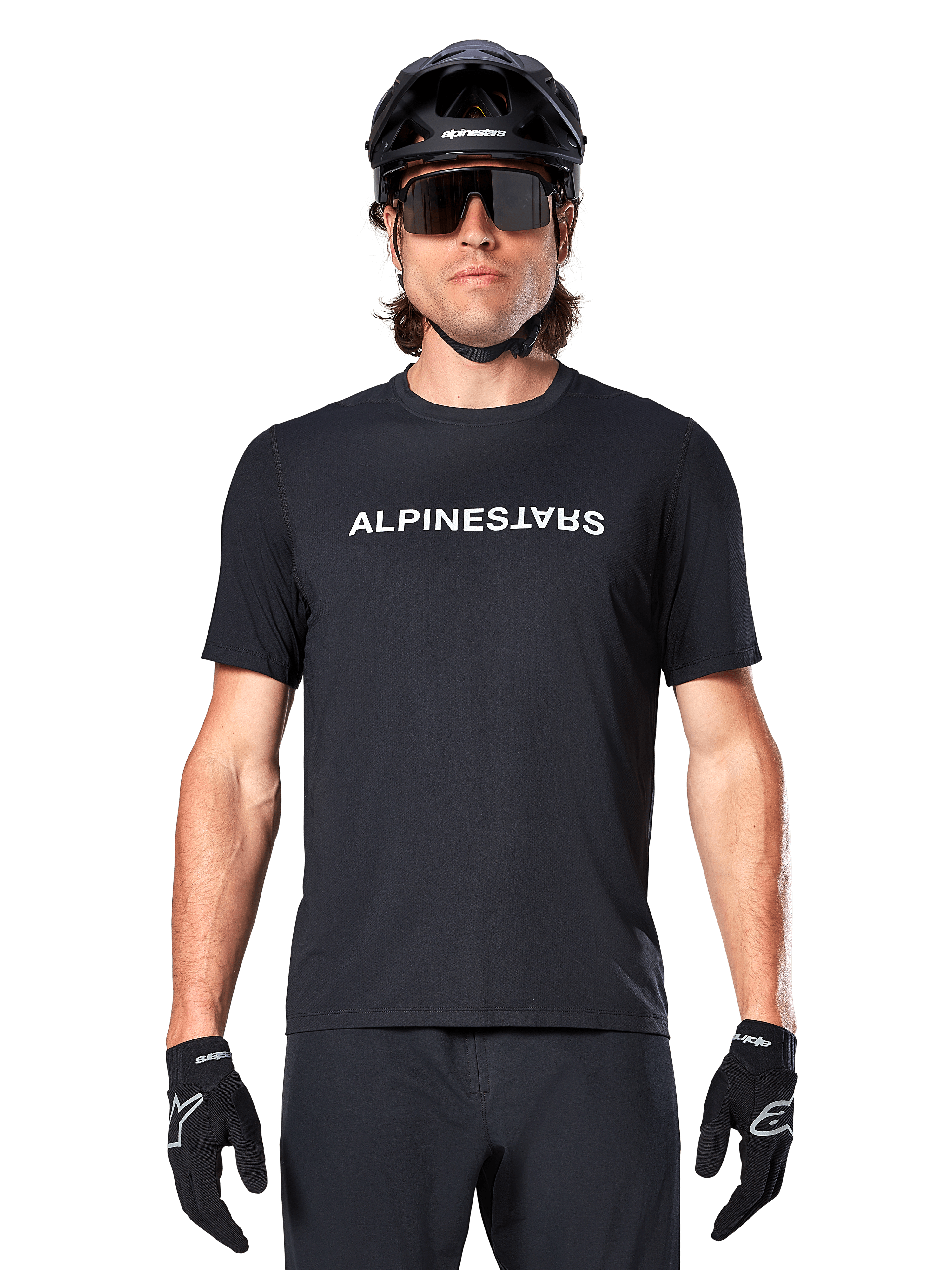 Mountain bike rider wearing the Alpinestars A-Dura Switch Short Sleeve Jersey in Black, featuring a white wordmark logo on the chest, styled with a black helmet, large sunglasses, and black gloves, professional cycling apparel for trail riding.