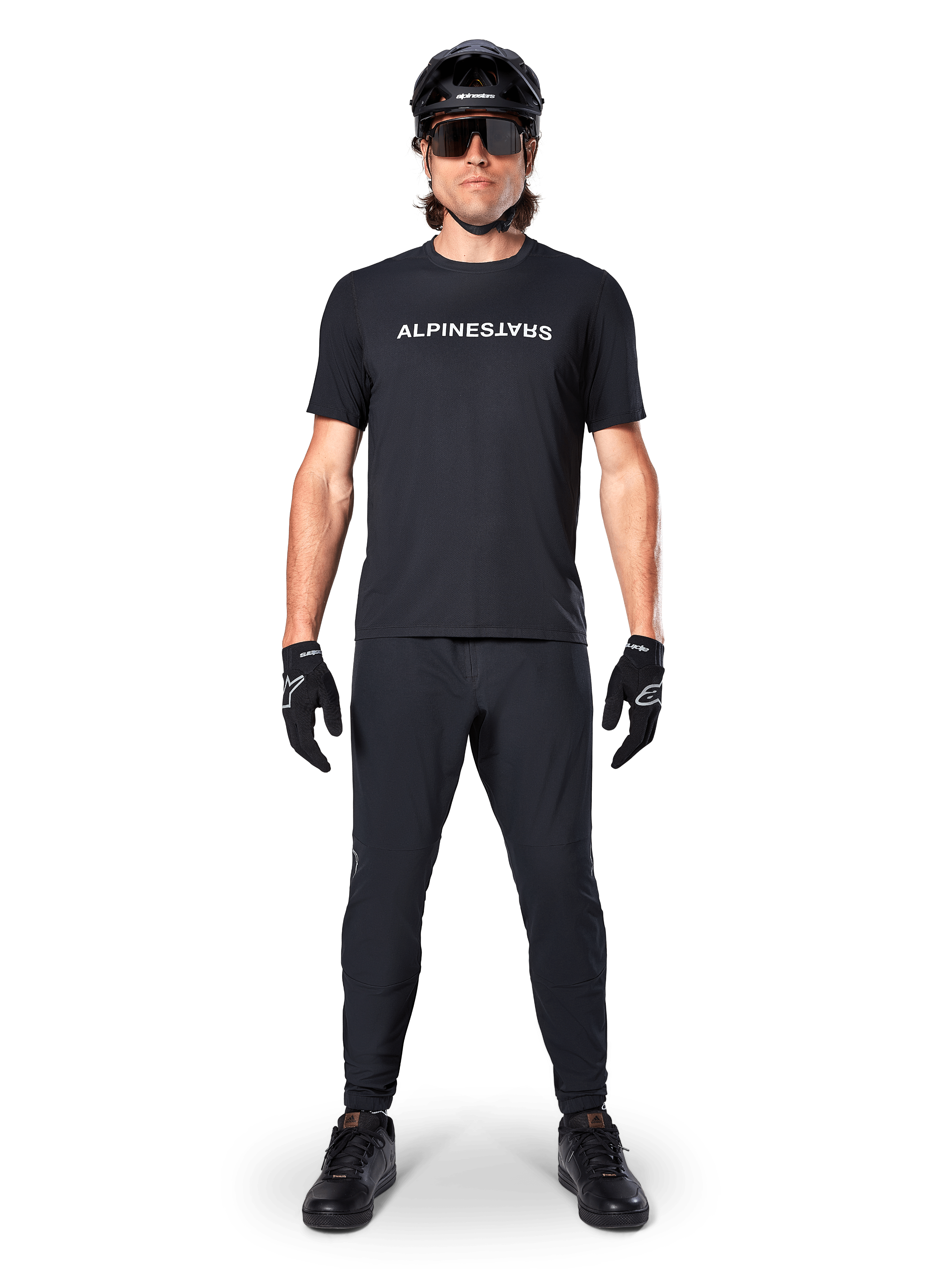 Mountain bike rider wearing the Alpinestars A-Dura Switch Short Sleeve Jersey in Black, featuring a white stylized wordmark logo on the chest, paired with matching black MTB pants, helmet, and gloves, complete trail-ready cycling outfit.
