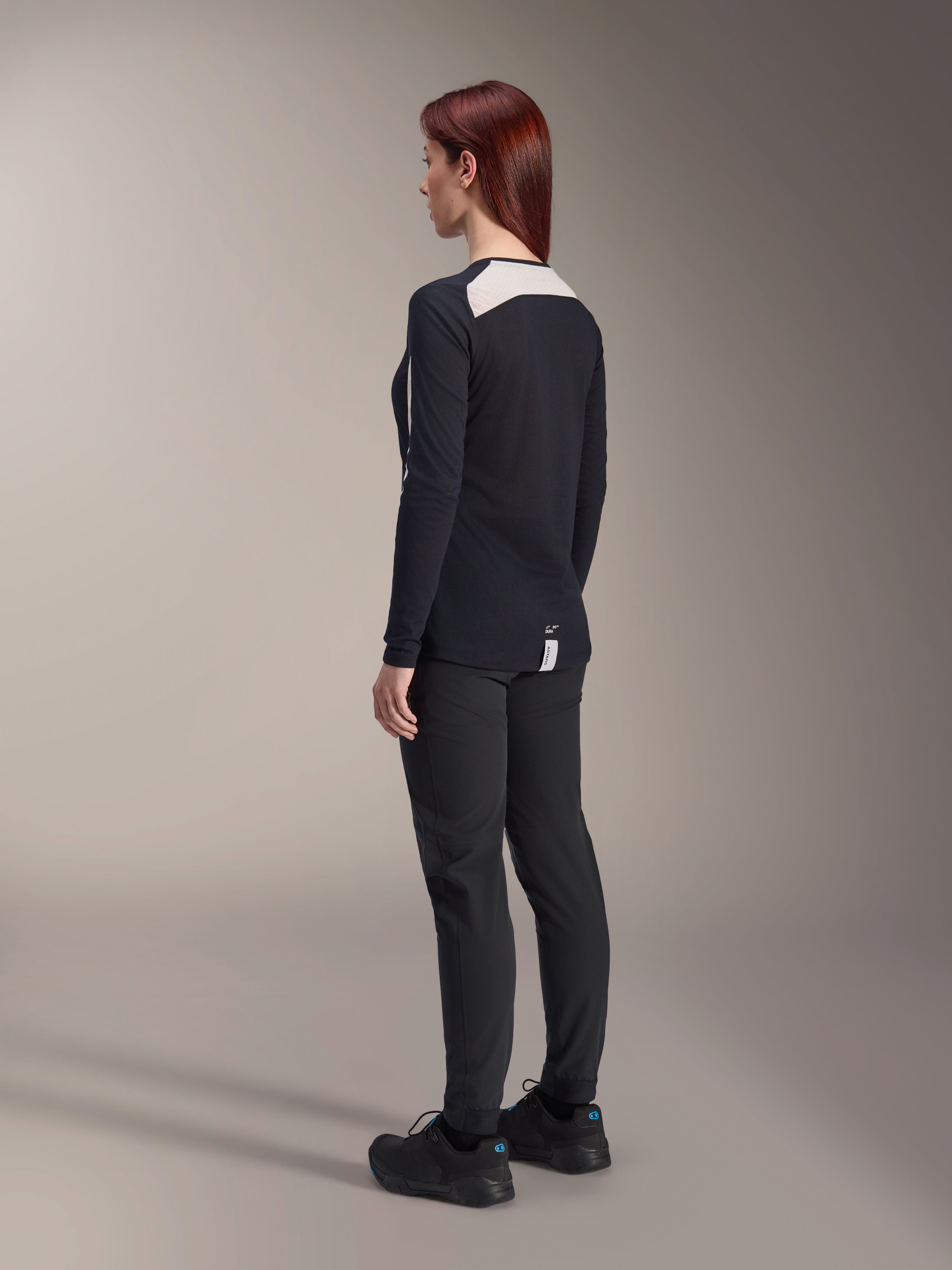 Female rider wearing Alpinestars Women Stella A-Dura Pants and Stella A-Dura Elite Jersey, Black, back view showing white mesh shoulder panels on long-sleeve jersey, slim-fit technical pants, coordinated performance mountain bike gear.