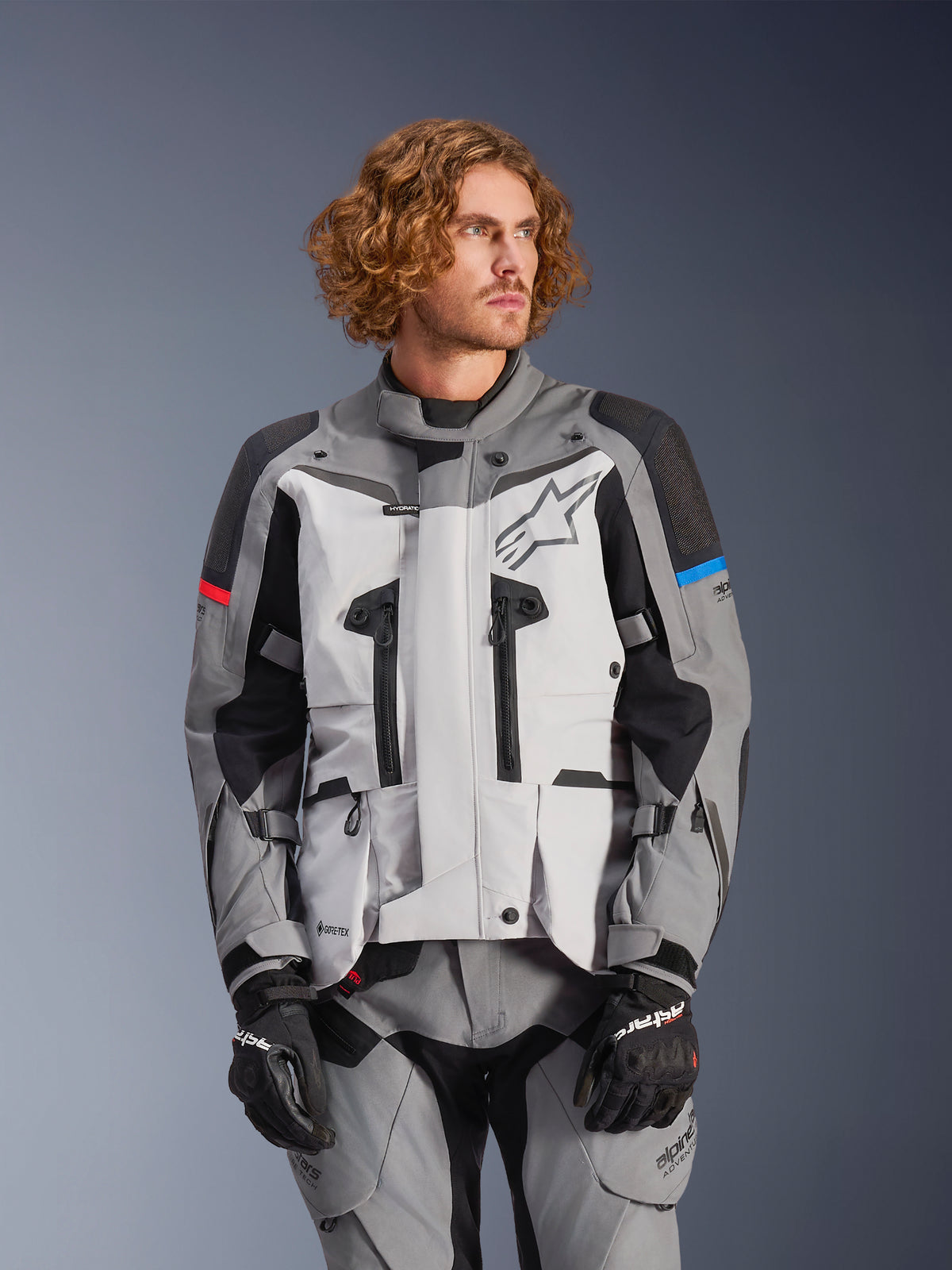 Alpinestars Boulder GORE-TEX Jacket, Motorcycle Adventure Touring Jacket, Gray and Black with Red and Blue Accents, Waterproof and Breathable, Star Logo, High Collar, Multiple Zippers, Worn with Matching Gray Pants and Black Gloves