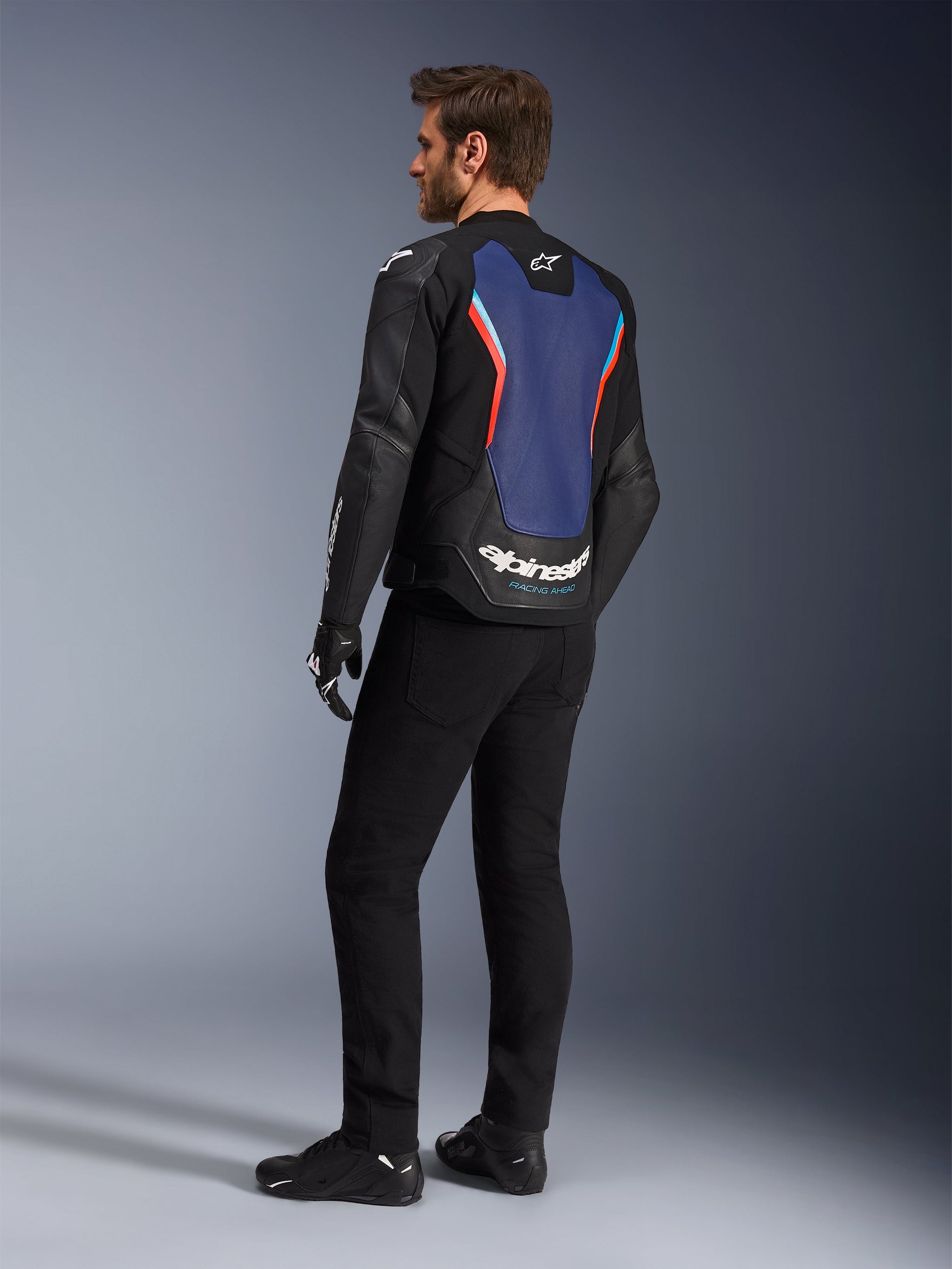 Rear three-quarter view of a male model wearing the Alpinestars GP Force V2 Leather Jacket in Black Blue Red Fluo, highlighting the blue back panel with red fluorescent accents and white branding, styled with black pants and motorcycle gloves.