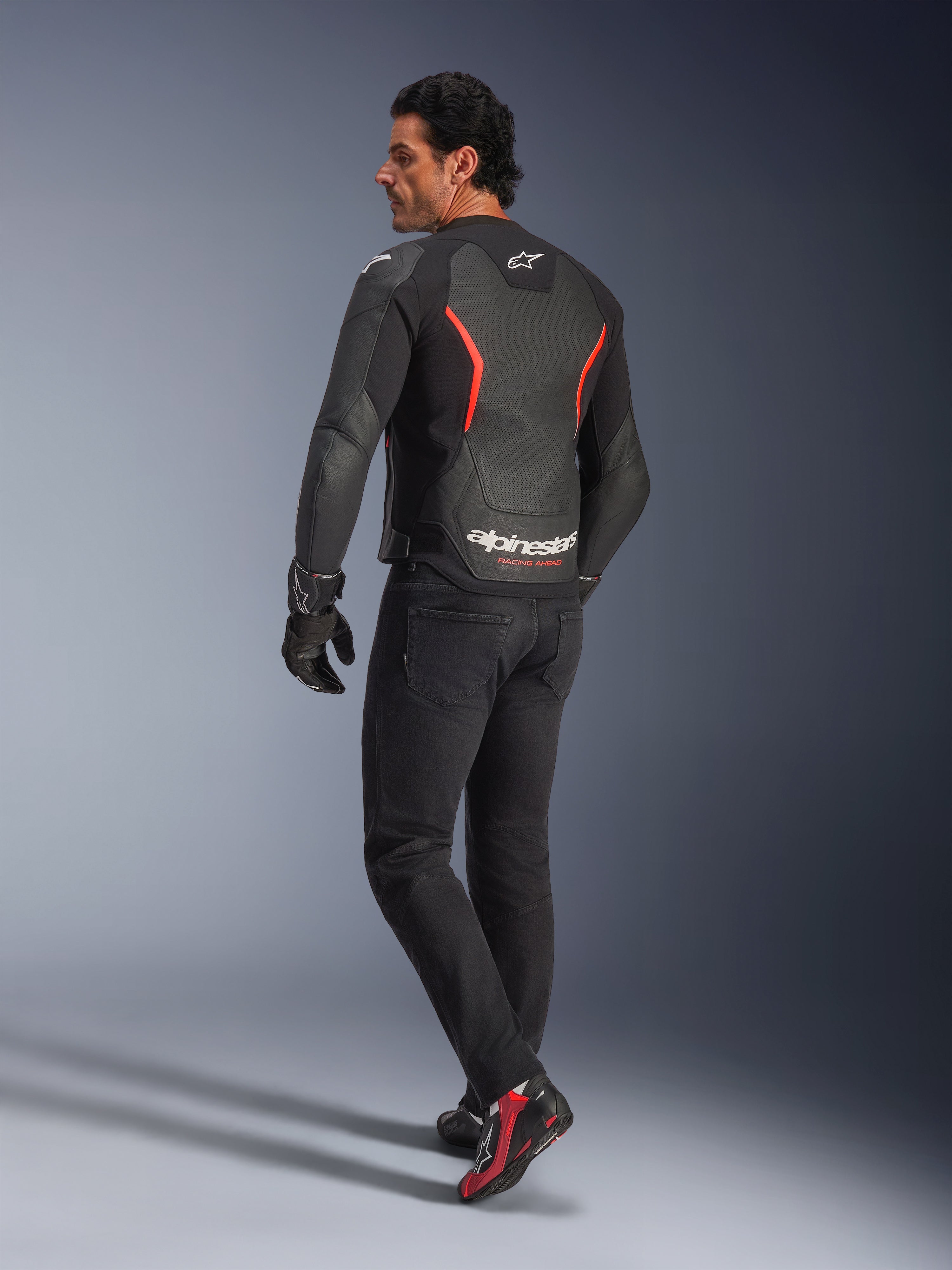 Male model from the back wearing the Alpinestars GP Force V2 Air Leather Jacket in Black Red Fluo, featuring extensive back perforations and bold white branding, paired with black riding jeans, gloves, and black and red riding boots in a studio setting.