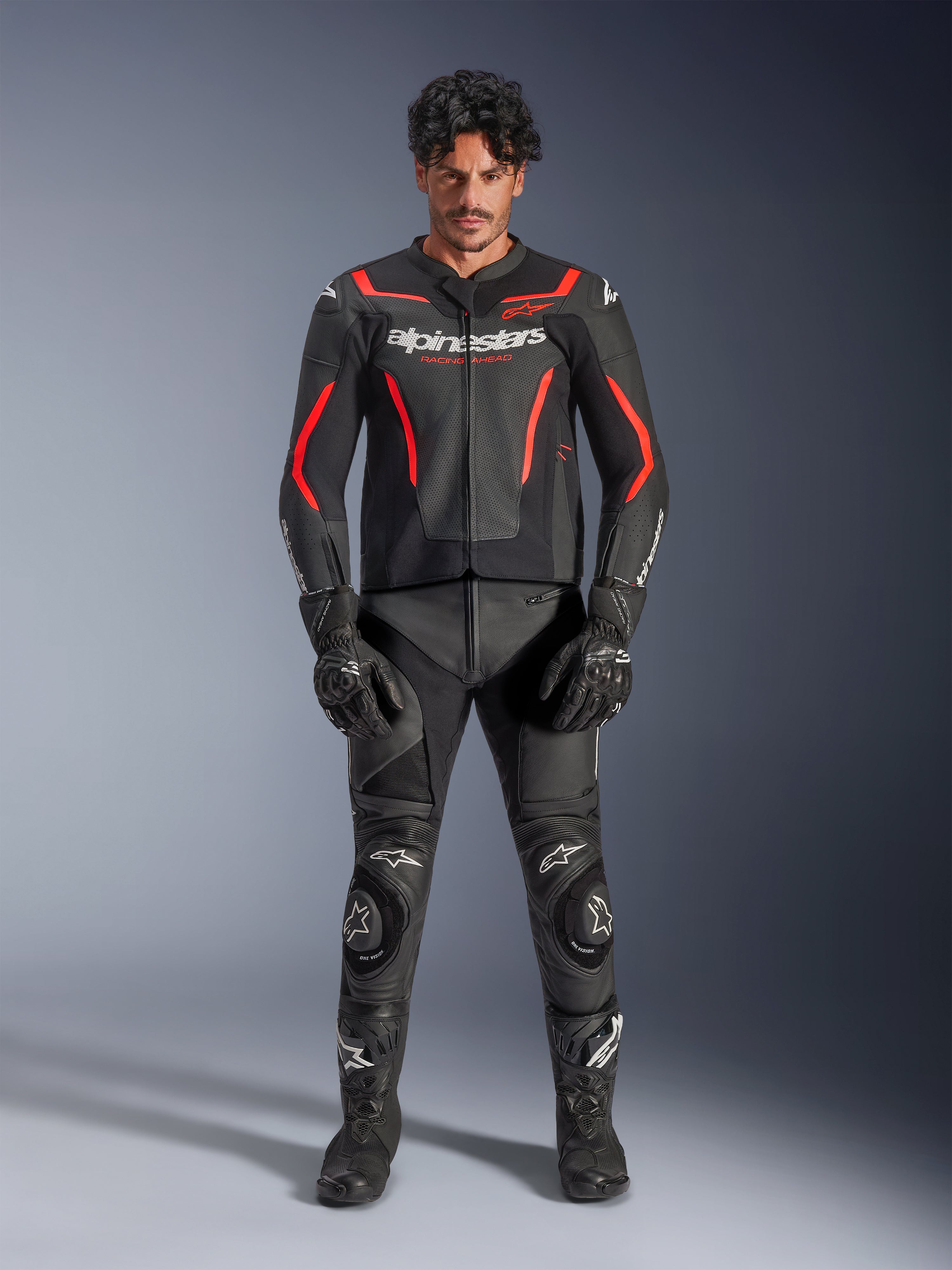 Male model wearing Alpinestars GP Force V2 Air Leather Jacket in Black and Red Fluo, featuring perforated chest panels and white branding, paired with black motorcycle leather pants, black gloves, and black boots for a complete racing look.