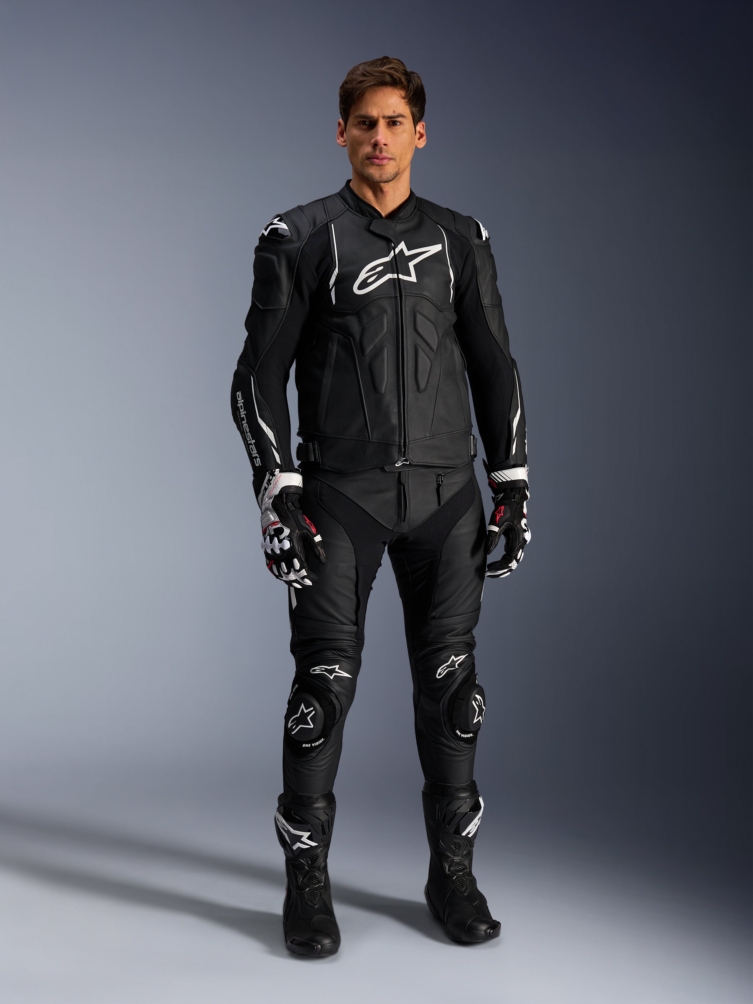 Male model wearing the Alpinestars Dusk Leather Jacket in Black and White, featuring prominent white logos on the chest and shoulders, paired with matching black leather motorcycle pants, black and white gloves, and black boots, complete sport racing outfit for road use.