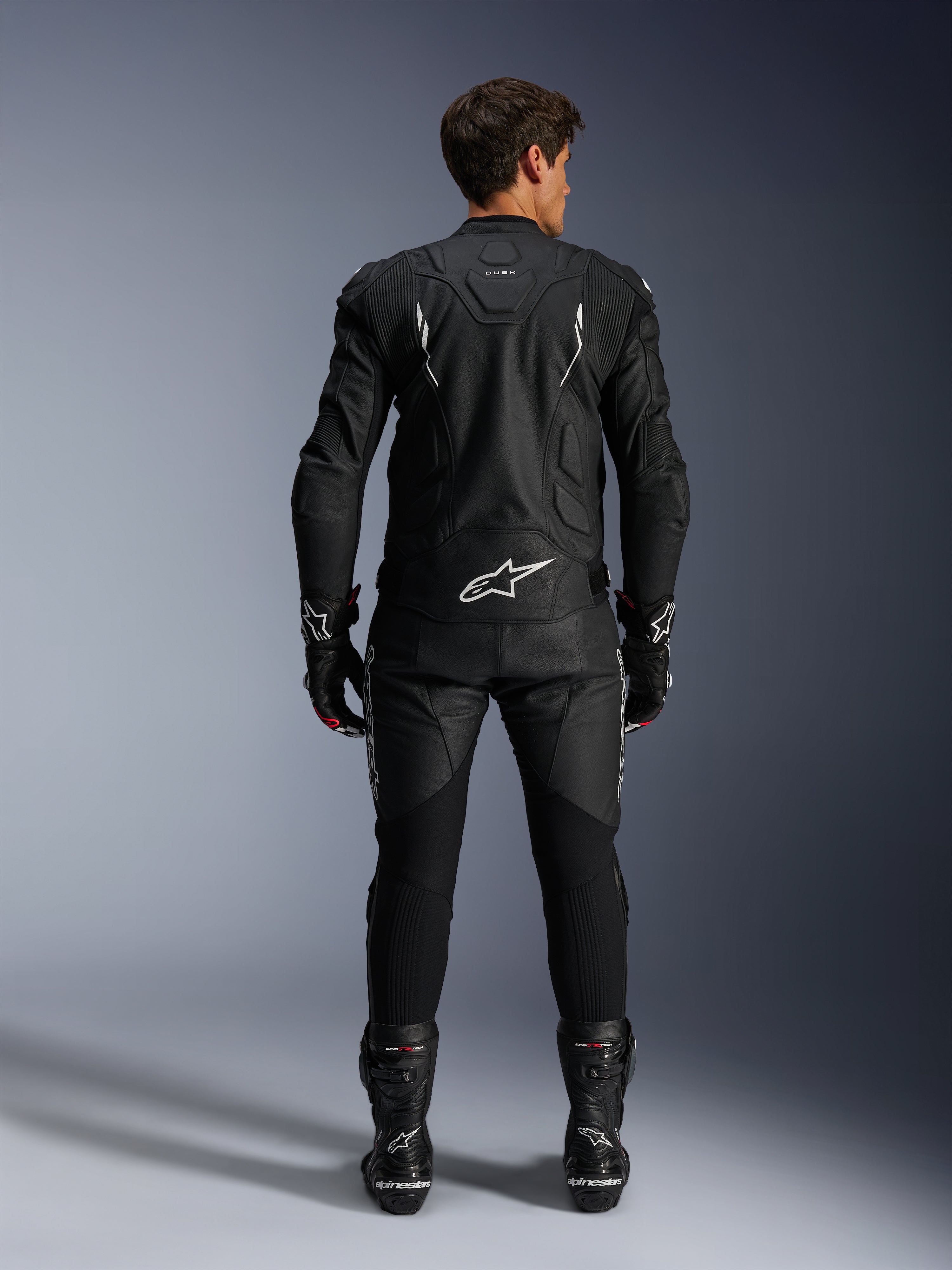 Motorcycle rider from a rear view wearing the Alpinestars Dusk Leather Jacket and Missile V3 Leather Pants, full black leather suit with white accents, jacket features 'Dusk' branding and Alpinestars logo on the back, paired with black racing boots and gloves, coordinated motorsport outfit for road racing.