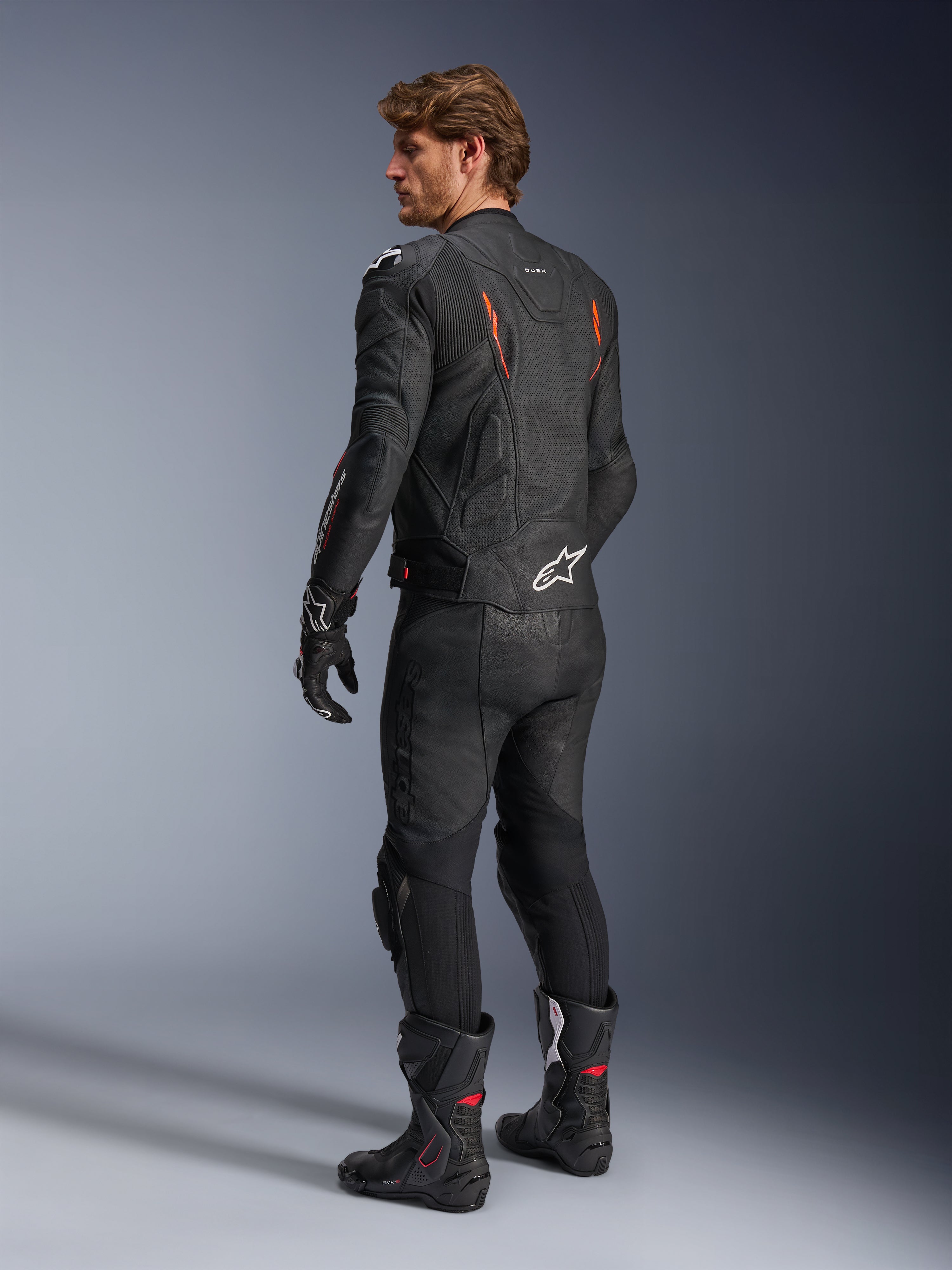 Alpinestars Dusk Airflow Leather Jacket, Black with Red Fluo accents, rear view of a rider in full leather motorcycle gear including matching black pants, gloves, and racing boots, featuring perforated leather and white Alpinestars logos.
