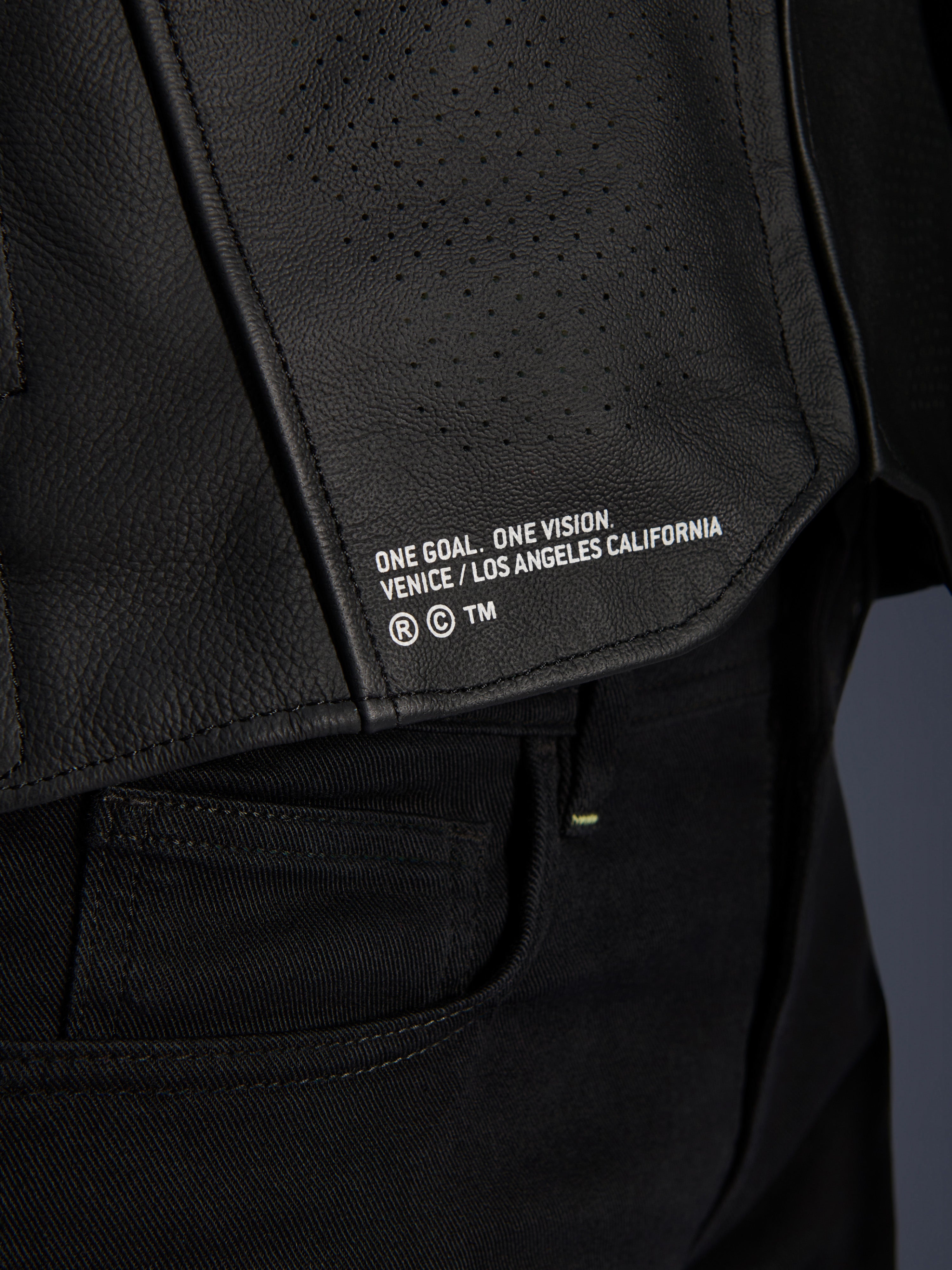 Close-up of the Alpinestars Atem V5 Leather Jacket in Black, featuring the lower back panel with 'ONE GOAL. ONE VISION. VENICE / LOS ANGELES CALIFORNIA' branding and perforated leather texture.