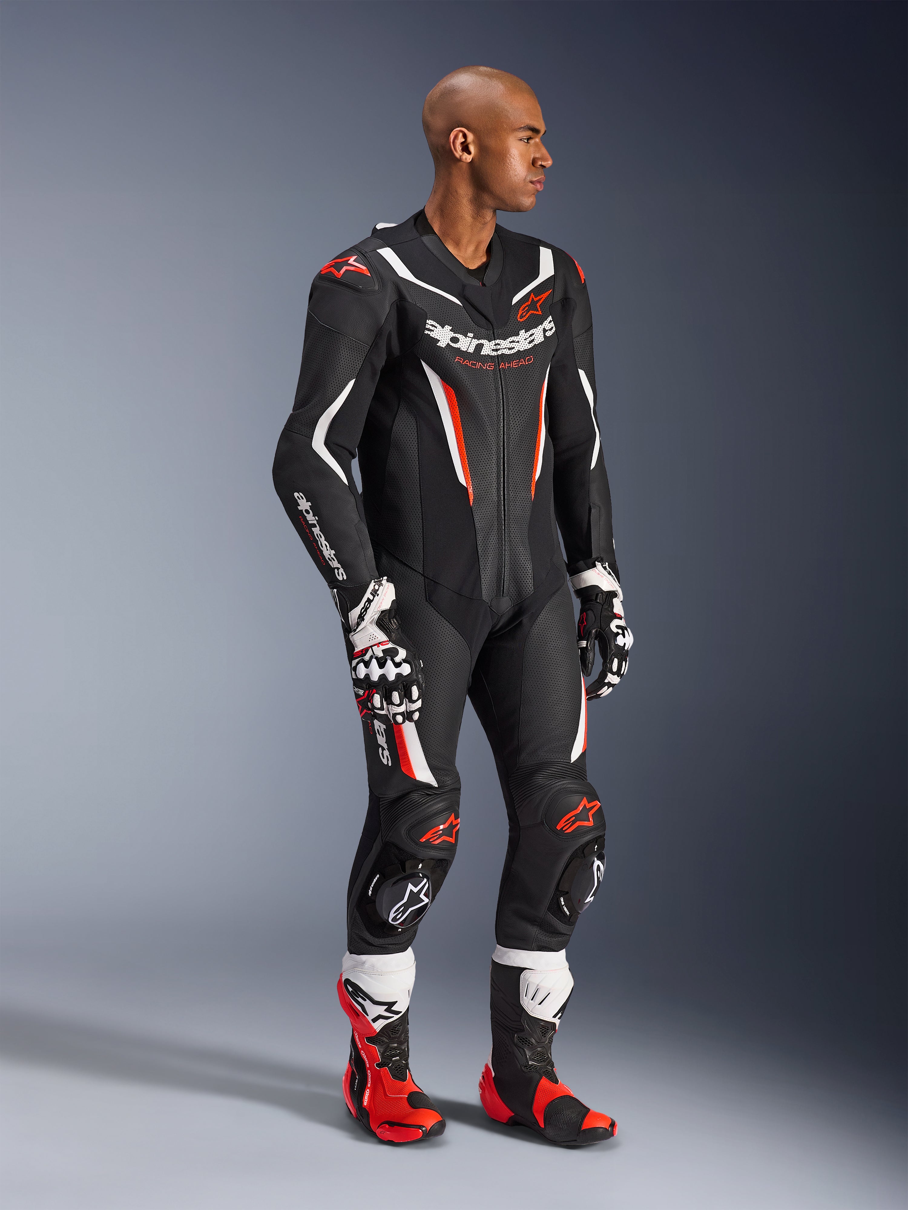 Model wearing the Alpinestars GP Force V2 Leather Suit 1-Piece in Black White Red Fluo, high-performance racing gear with perforated leather panels and DFS Lite shields, styled with matching red and white racing boots and gloves, professional motorsport aesthetic.